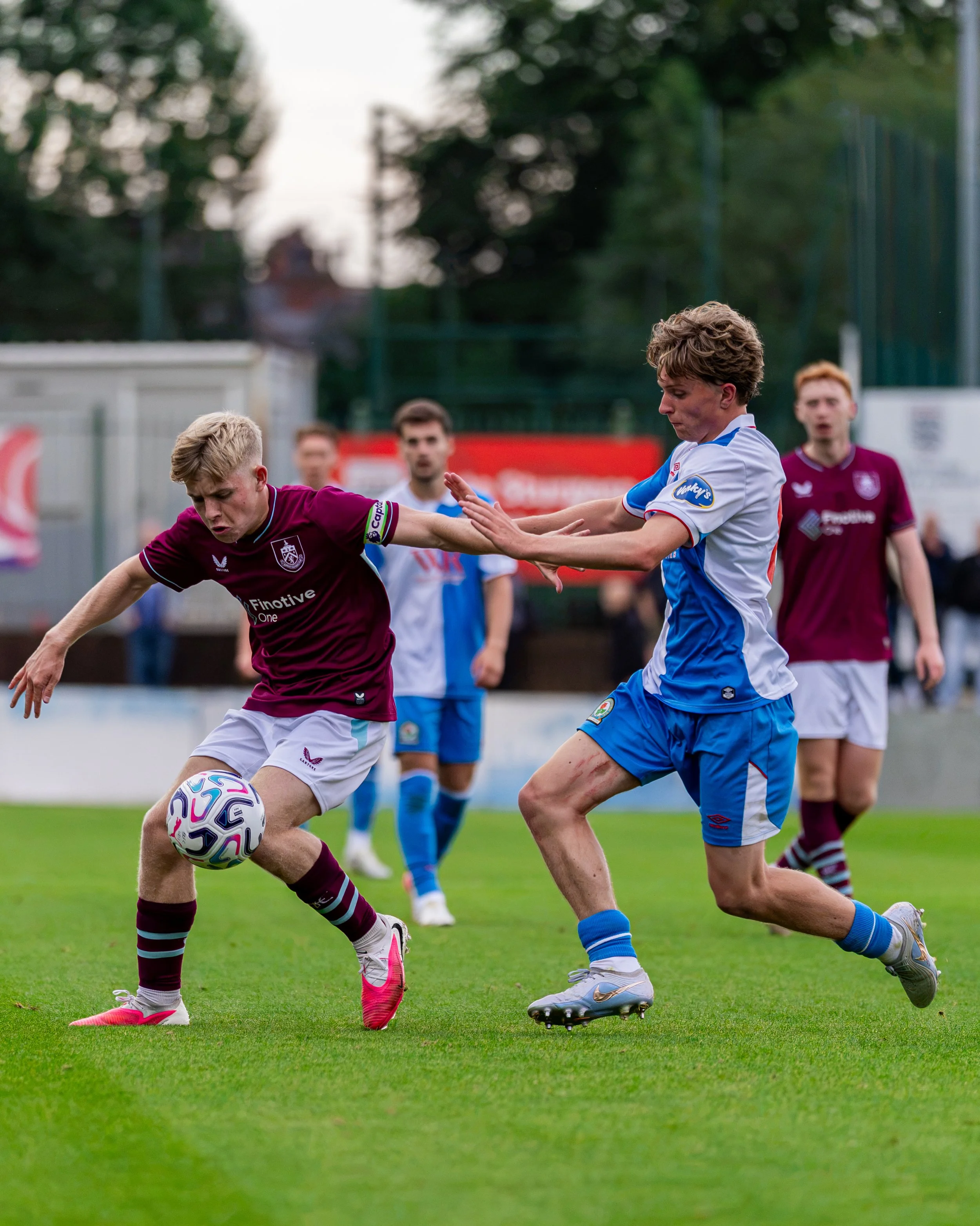 Two soccer players are competing for possession of the ball on the field. One player is wearing a maroon jersey with white shorts and pink cleats, and the other is wearing a blue and white jersey with blue shorts and gray cleats. Several other player