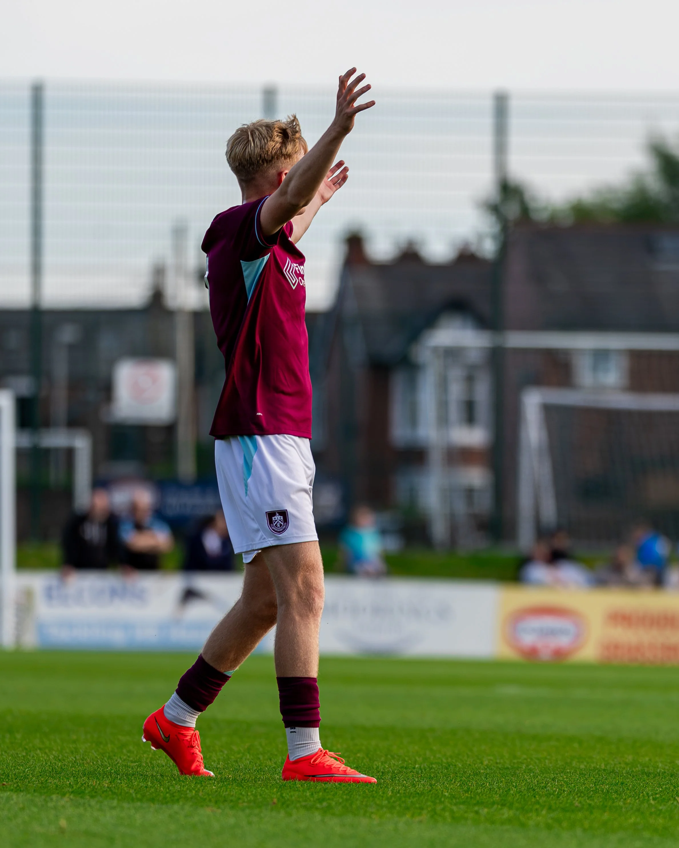 A soccer player in a maroon jersey, white shorts, and red cleats is standing on a grass field, raising his arms with open hands during a game.