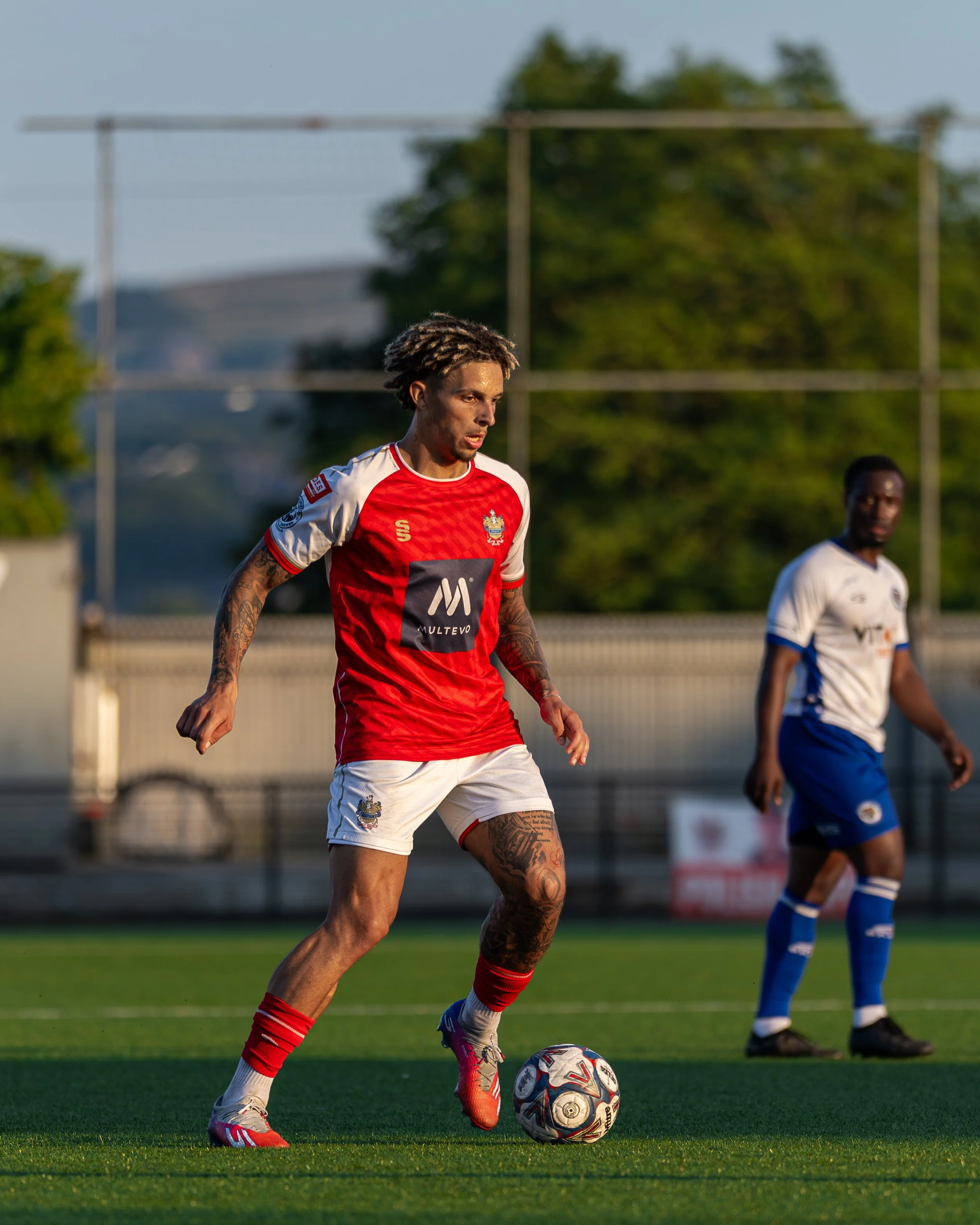 A soccer player with dreadlocks, tattoos, and a red and white uniform is preparing to kick a soccer ball on a field during daylight. There is another player in a white and blue uniform in the background.