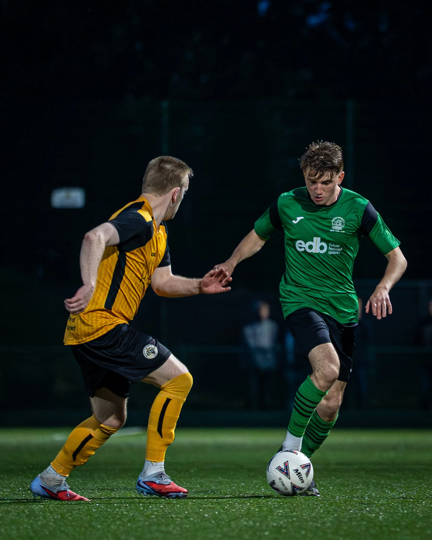 Two soccer players competing for the ball on a field during a night game. One player wears a green jersey and black shorts, the other wears a black and yellow striped jersey and yellow socks.