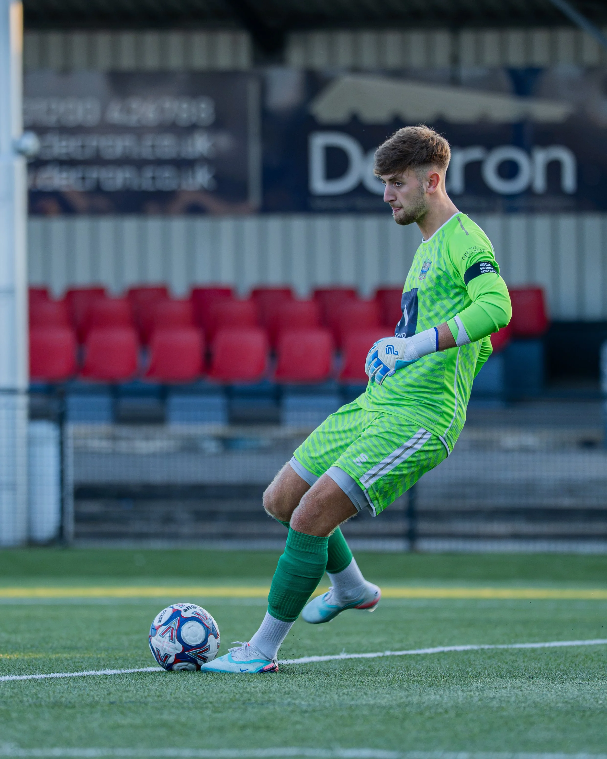 A soccer goalkeeper in a neon green jersey and gloves is about to kick a soccer ball on a green field during daytime.
