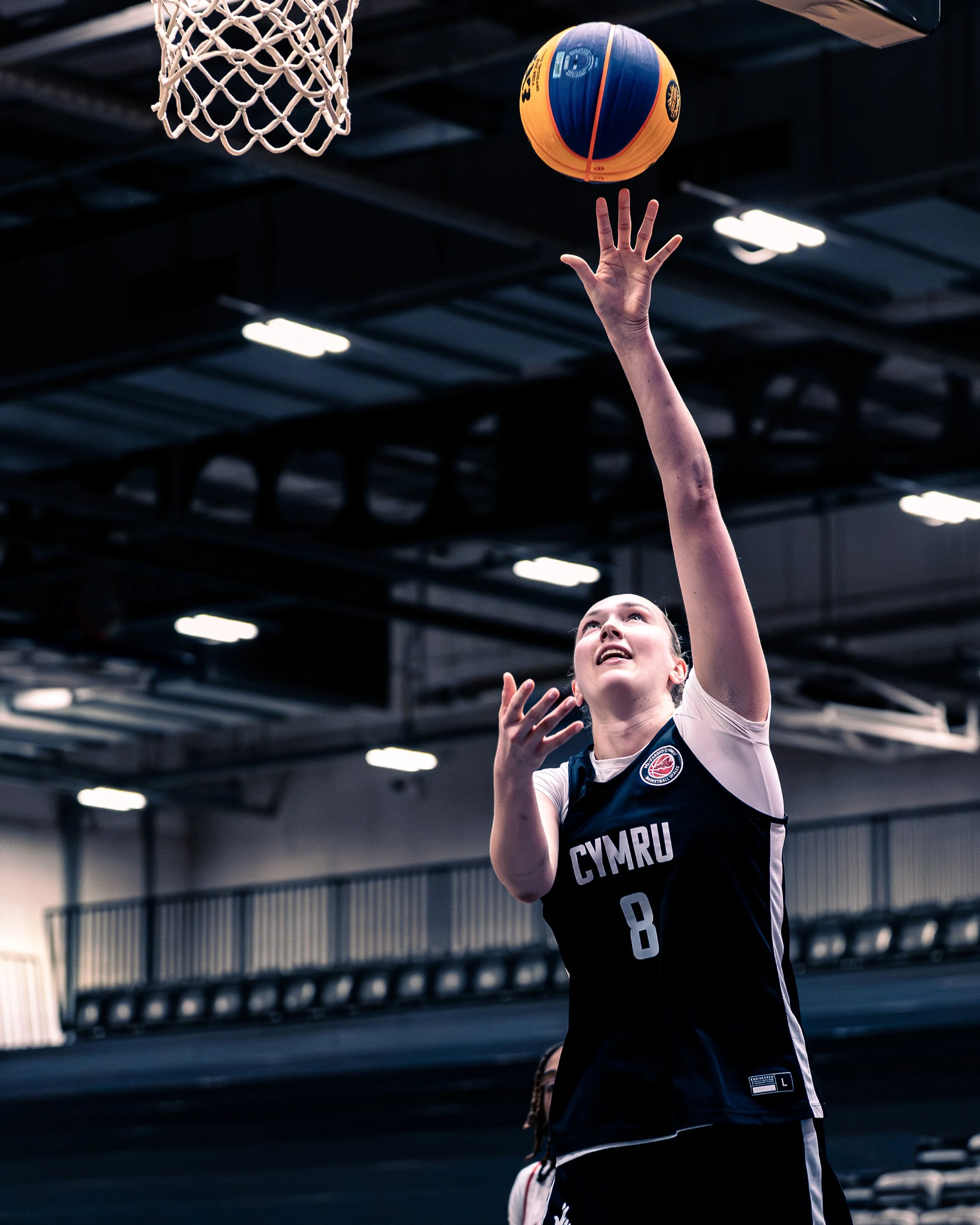 A female basketball player in a black jersey with the word 'CYMRU' on it, reaching up to shoot or grab a basketball near the hoop inside an indoor sports facility.