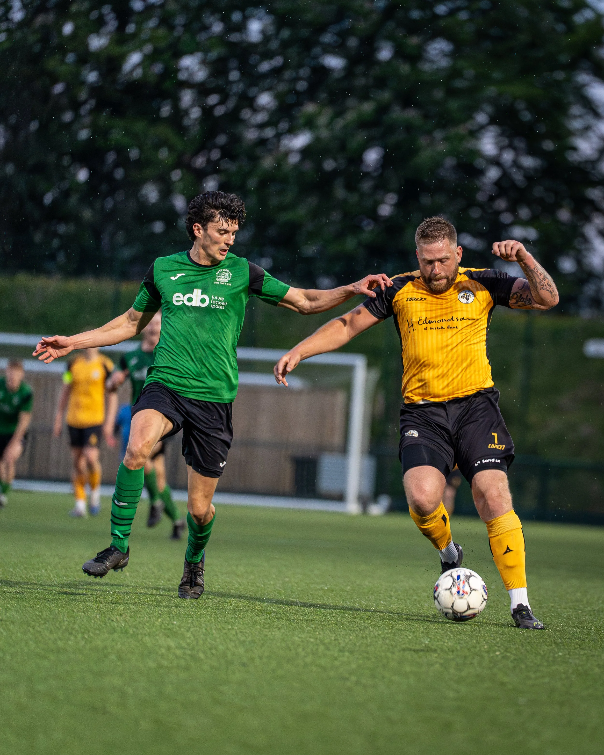 Two soccer players, one in a green jersey and the other in a yellow and black jersey, competing for the ball on a grass field during a match, with trees in the background.