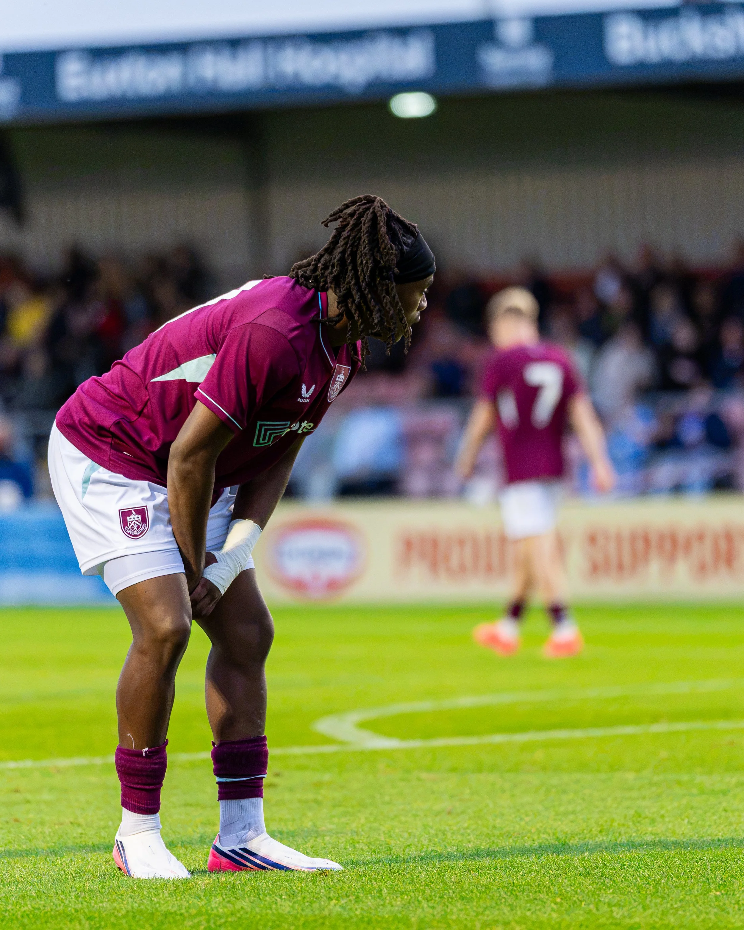 A female soccer player with long braided hair is bending forward on a green field, wearing a maroon or dark red jersey, white shorts, and white soccer cleats, with a crowd and stadium in the background.