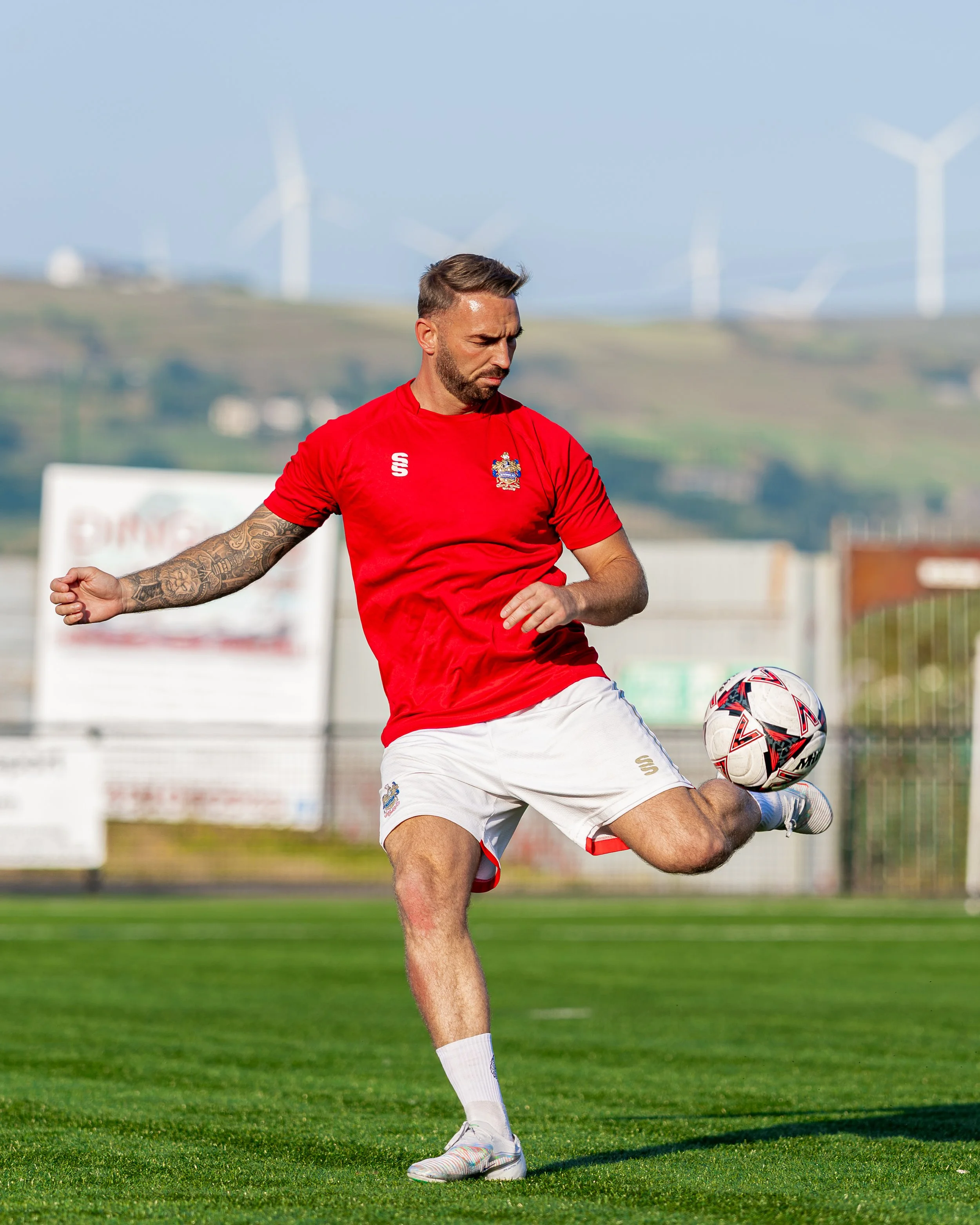 A man playing soccer outdoors on a grass field with wind turbines in the background. He is wearing a red shirt, white shorts, and colorful sneakers. He is in motion, kicking a soccer ball.