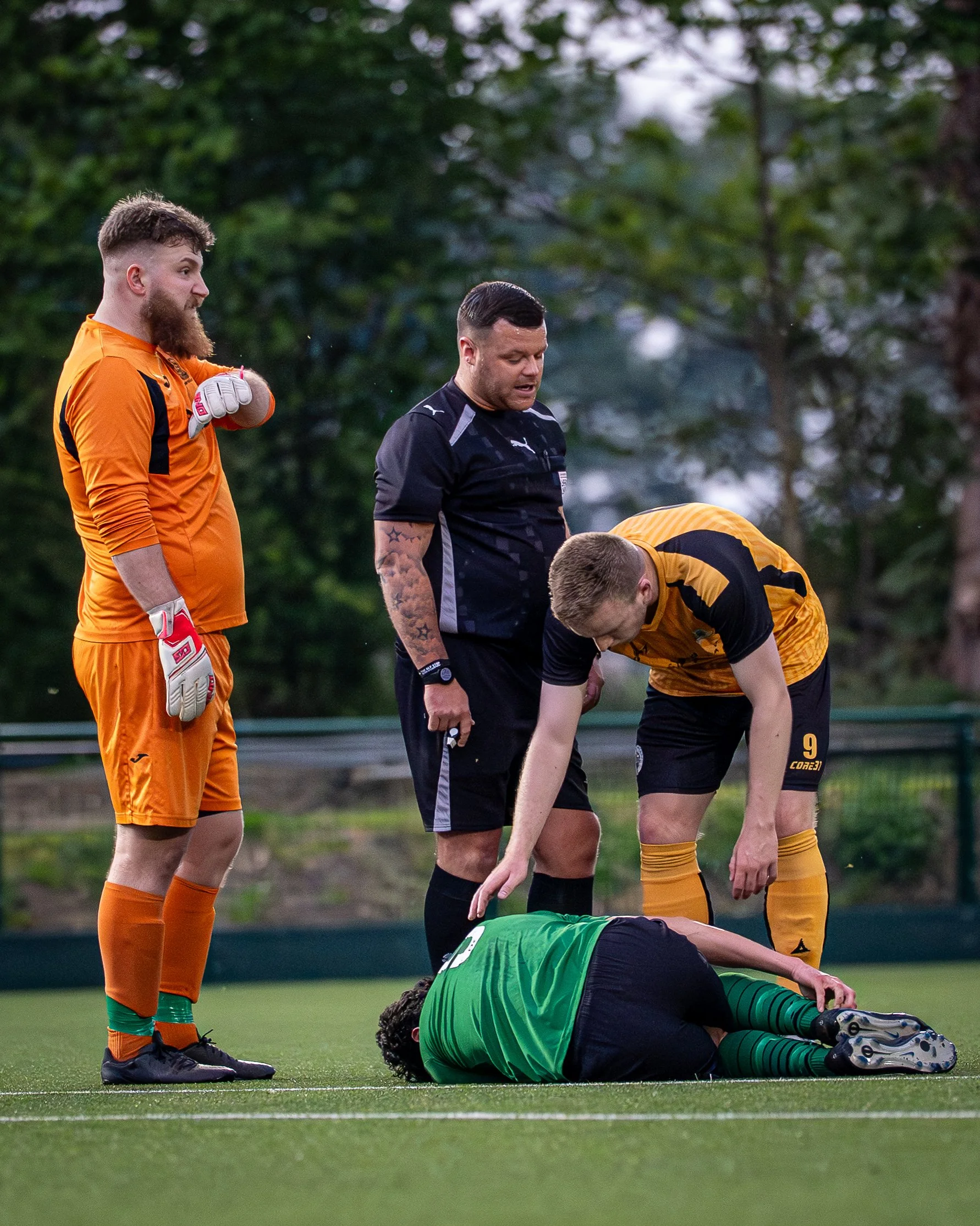 A soccer goalkeeper lying on the ground after a fall, with four other players and a referee standing nearby on the field. The goalkeeper wears a green jersey and black shorts, while the players are in yellow and black uniforms, and one player is in a