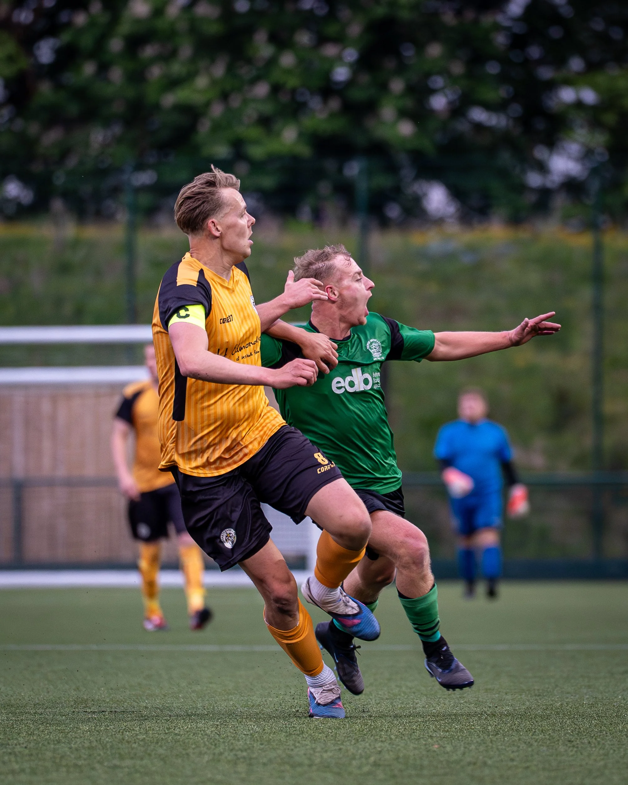Two soccer players, one in a yellow jersey and the other in a green jersey, competing for the ball on a field during a match, with a blurred background of other players and trees.