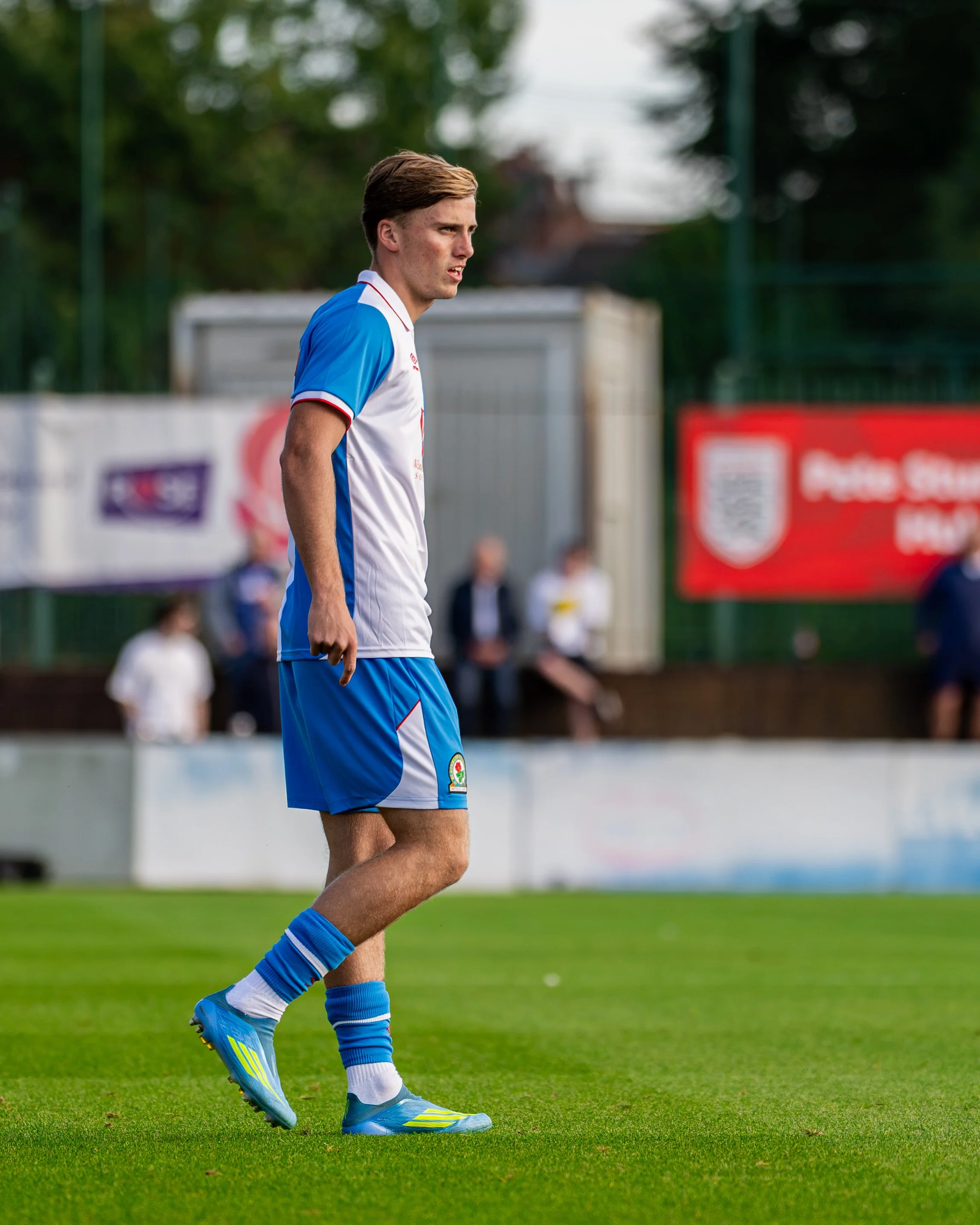 A young male soccer player wearing a white and blue uniform walking on a green soccer field, with blurred spectators and advertisements in the background.