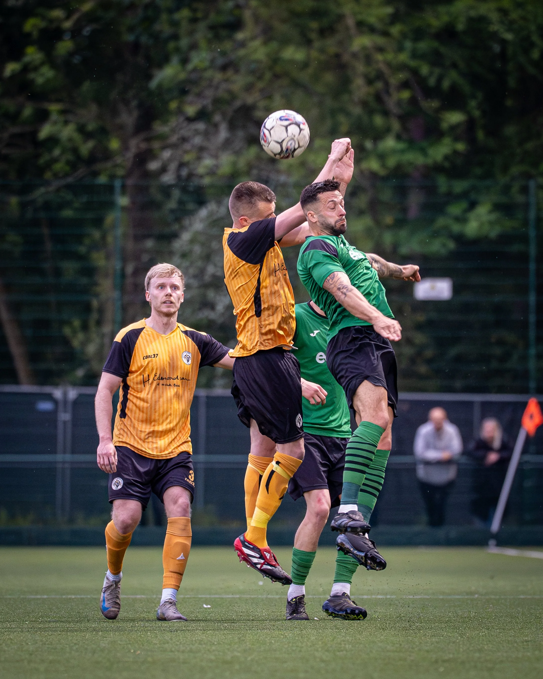 Two soccer players are jumping to head the ball during a match. One player is wearing a yellow and black uniform, and the other is in a green uniform. There is a third player in the background wearing a yellow uniform. The scene is set on a grassy fi