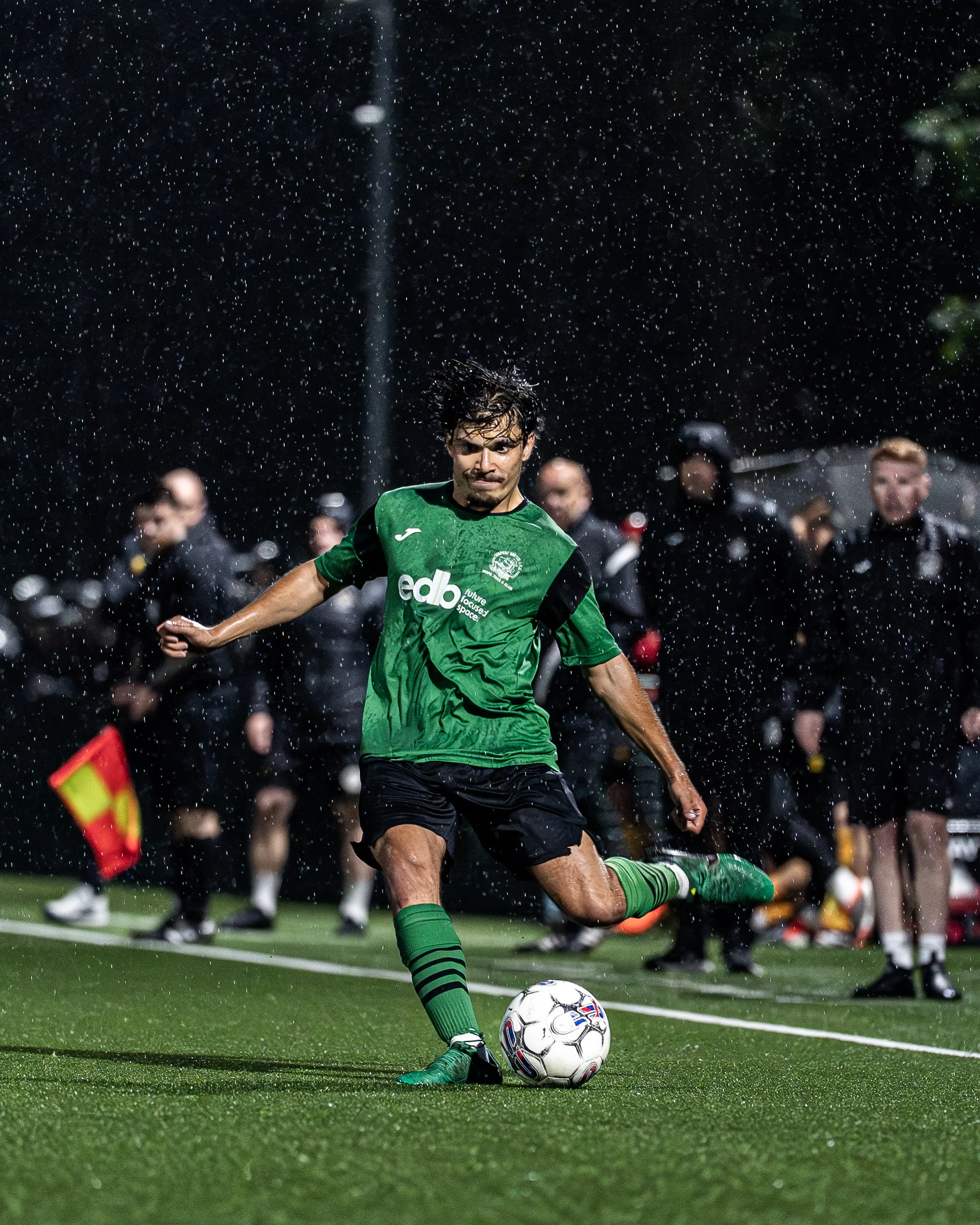 A soccer player in a green jersey kicks a ball during a match at night, with other players and spectators visible in the background.