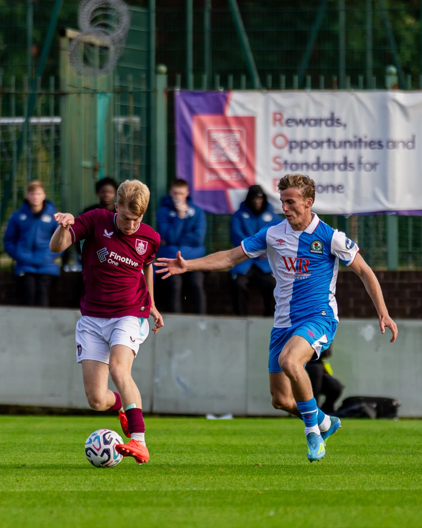 Two soccer players, one in a maroon jersey and the other in a blue and white jersey, competing for the ball on a green field with a fence and a banner in the background.