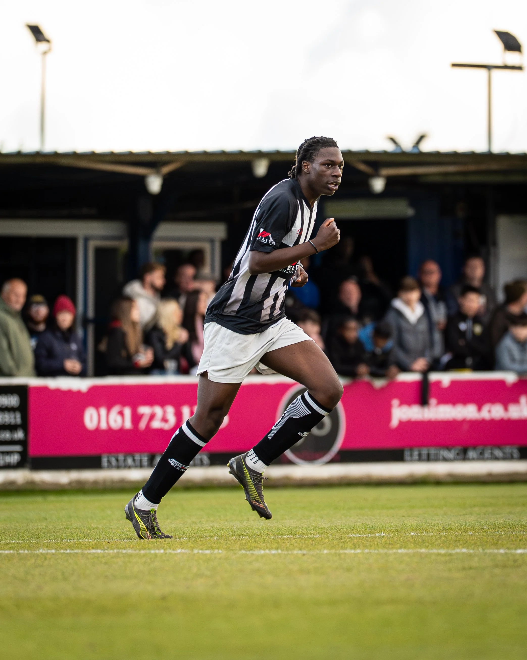 A soccer player running on the field during a game, wearing black and white striped jersey, white shorts, and black socks with stripes, with spectators watching from the stands in the background.