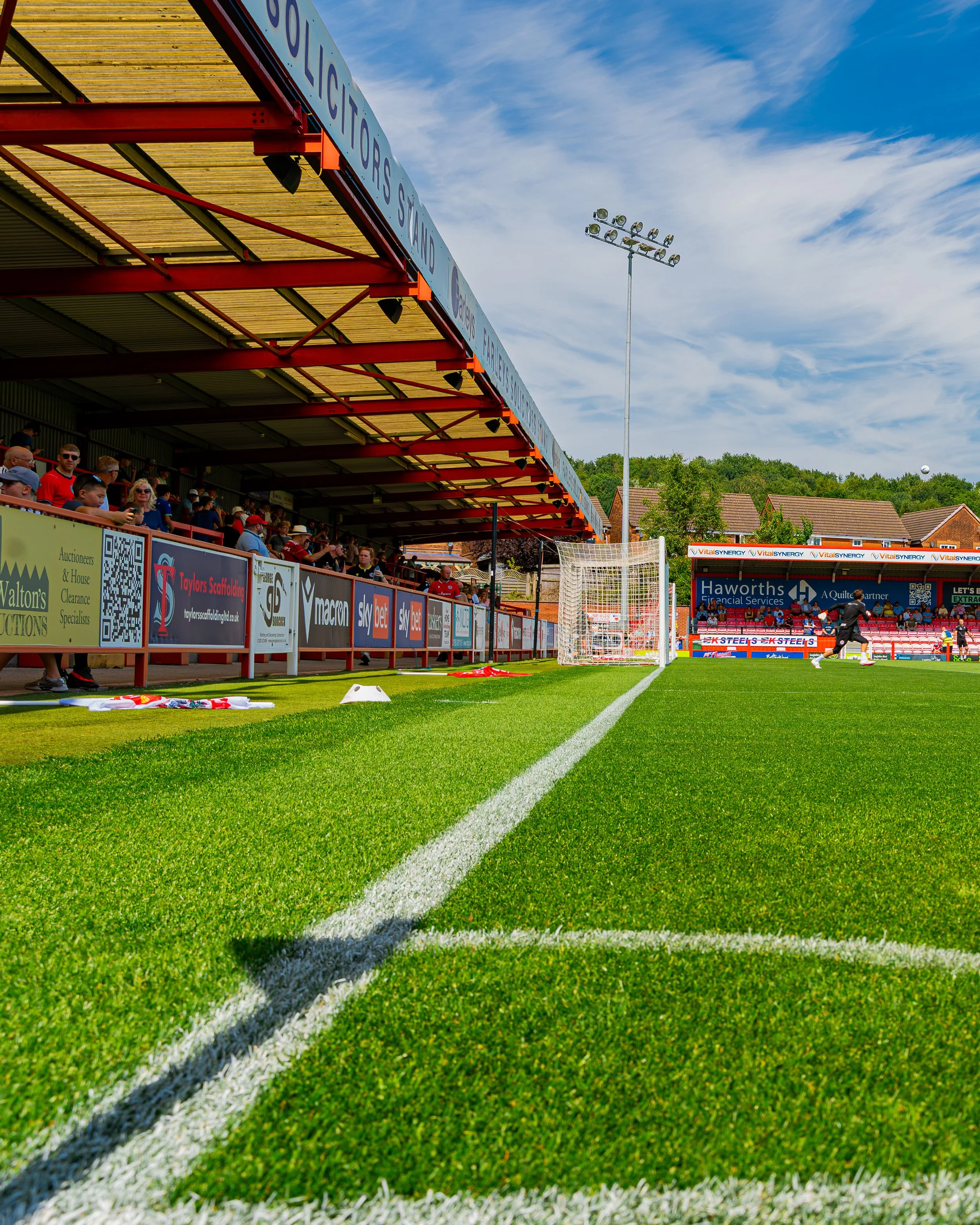 View of a football pitch with bright green grass, white boundary lines, and spectator stands with people, under a partly cloudy sky. There is a goalpost near the boundary, and floodlights are visible in the background.