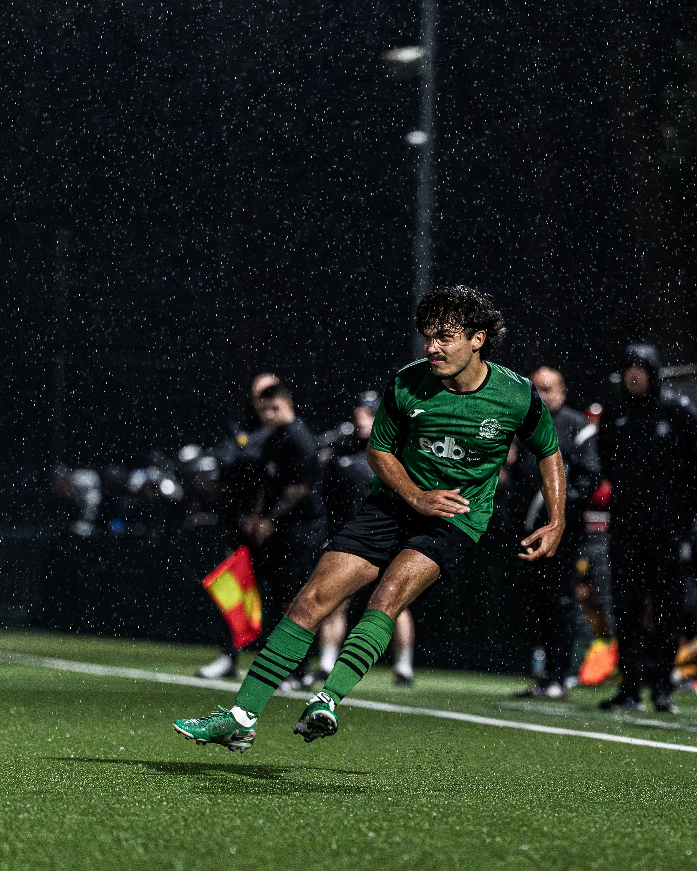 A soccer player in a green jersey and black shorts running on a wet soccer field at night, with rain falling around him.
