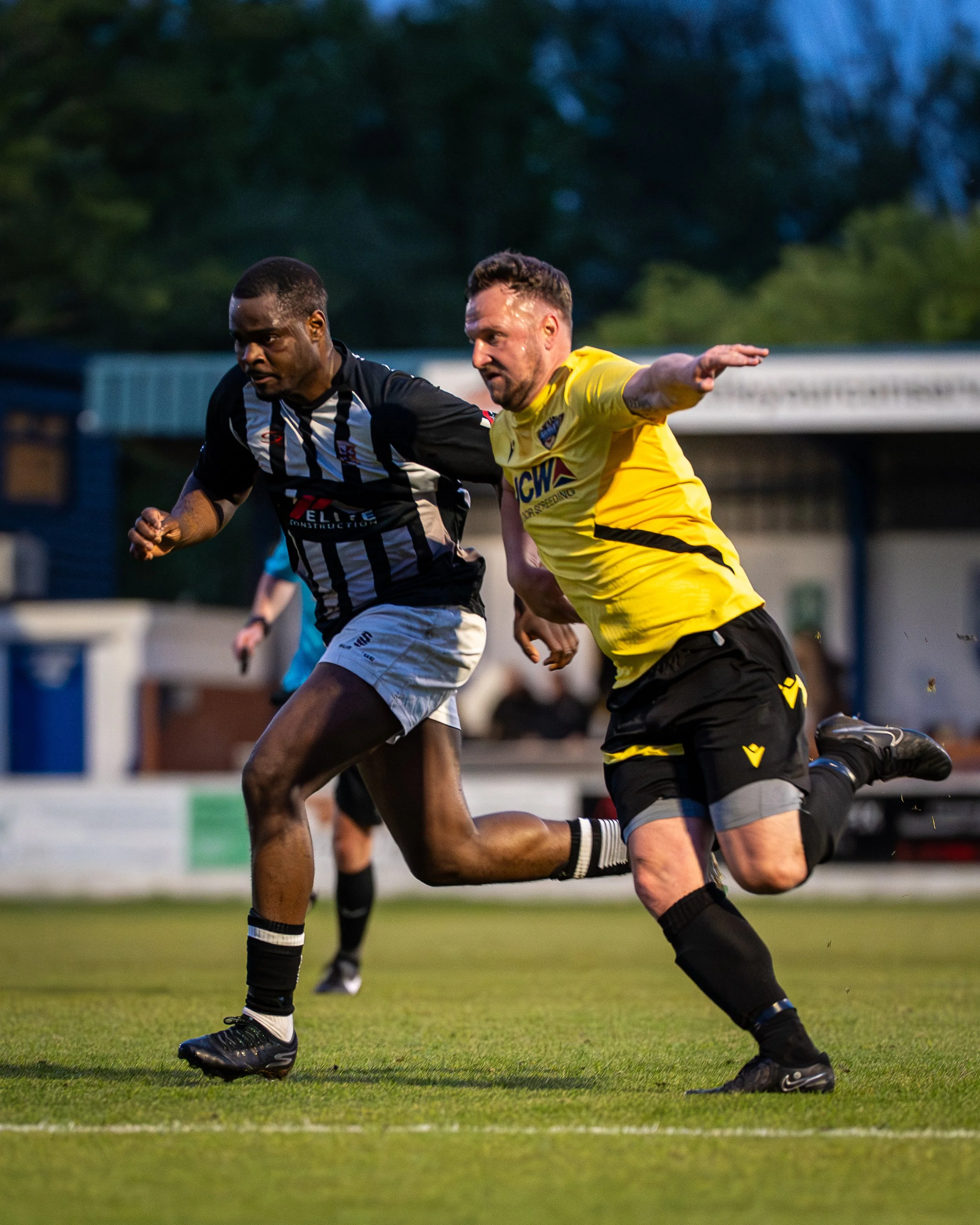 Two soccer players running on a field, one in a black and white striped jersey and the other in a yellow jersey, during a game in the evening.