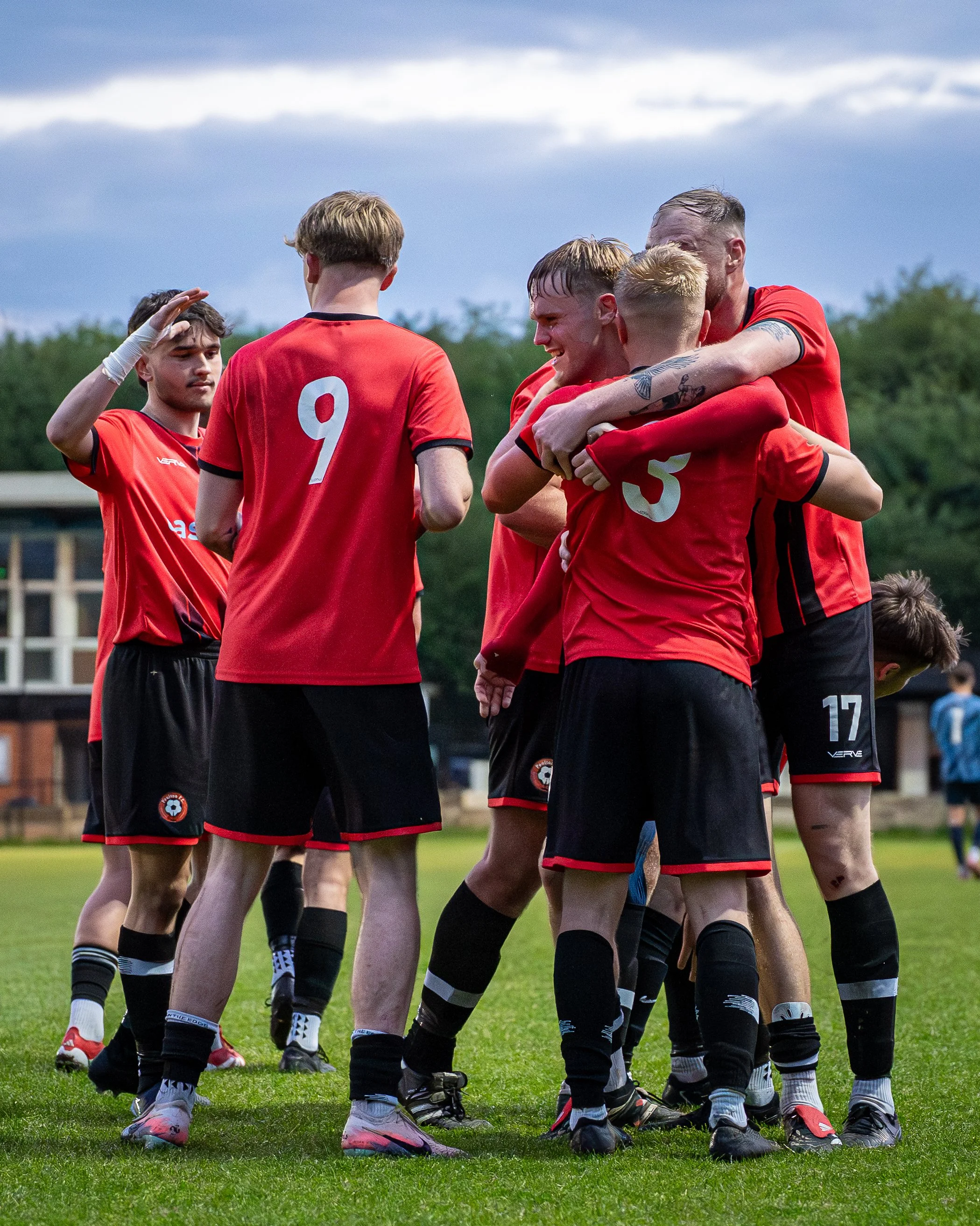 A group of male soccer players in red jerseys huddle together on a soccer field during a game or practice, some with arms around each other and one player saluting or shielding his eyes.