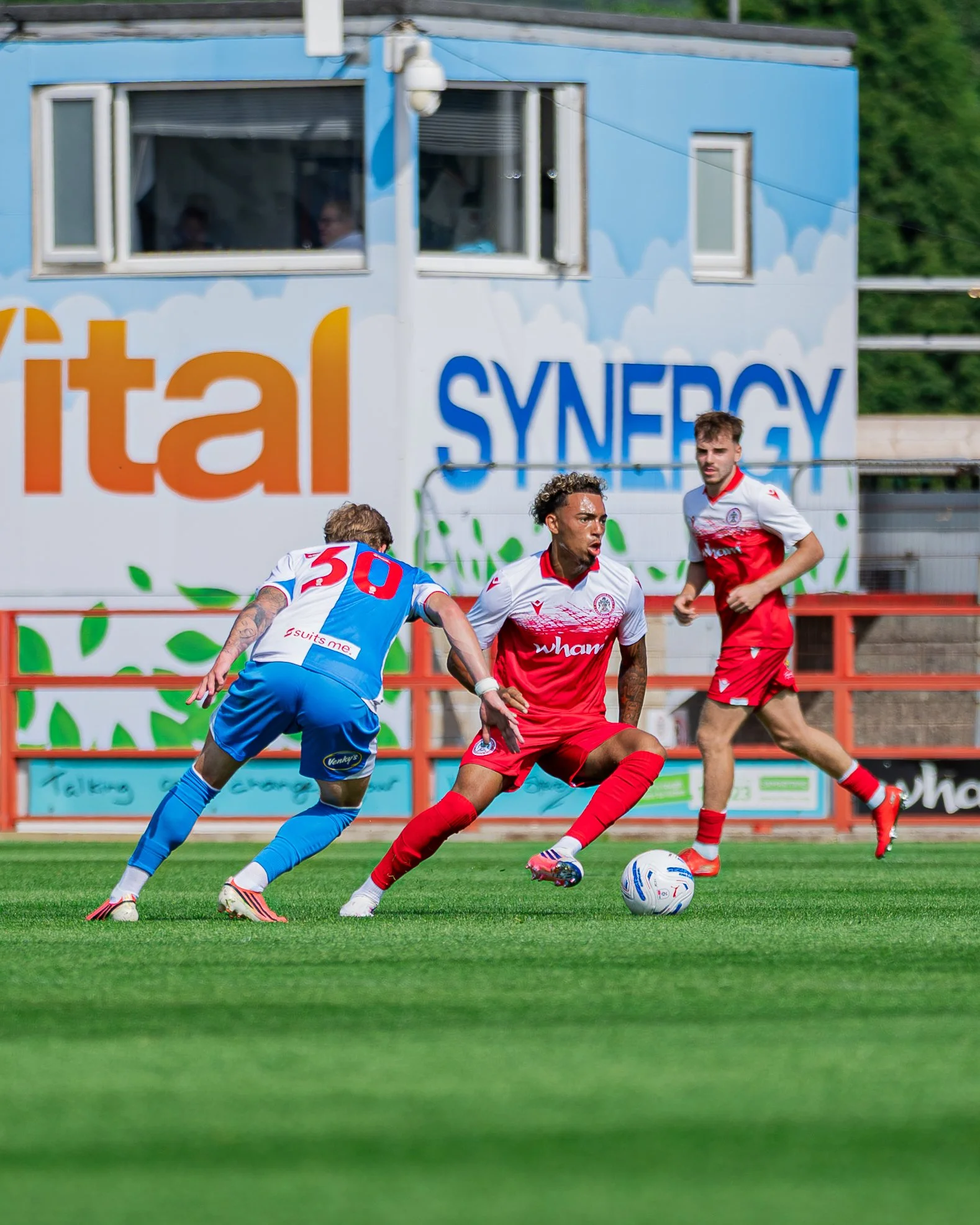 Soccer players in red and blue uniforms competing for the ball on a green field, with advertising banners in the background.