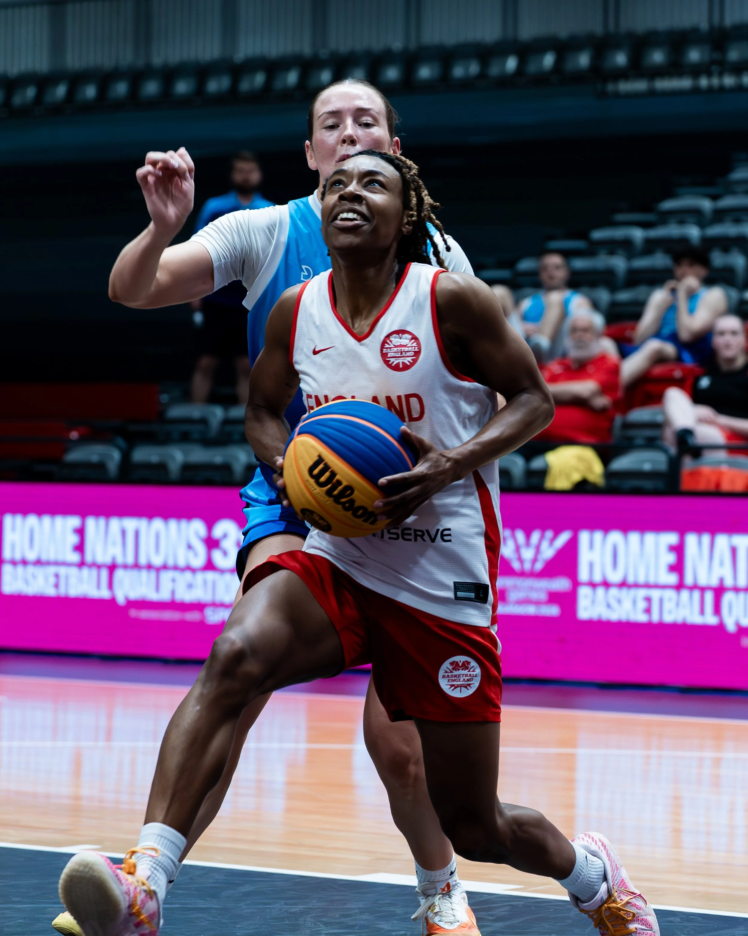 Two female basketball players are on the court during a game, with one player holding a basketball and the other closely defending. The player with the ball is wearing a white jersey with red accents and the player defending is wearing a blue jersey.