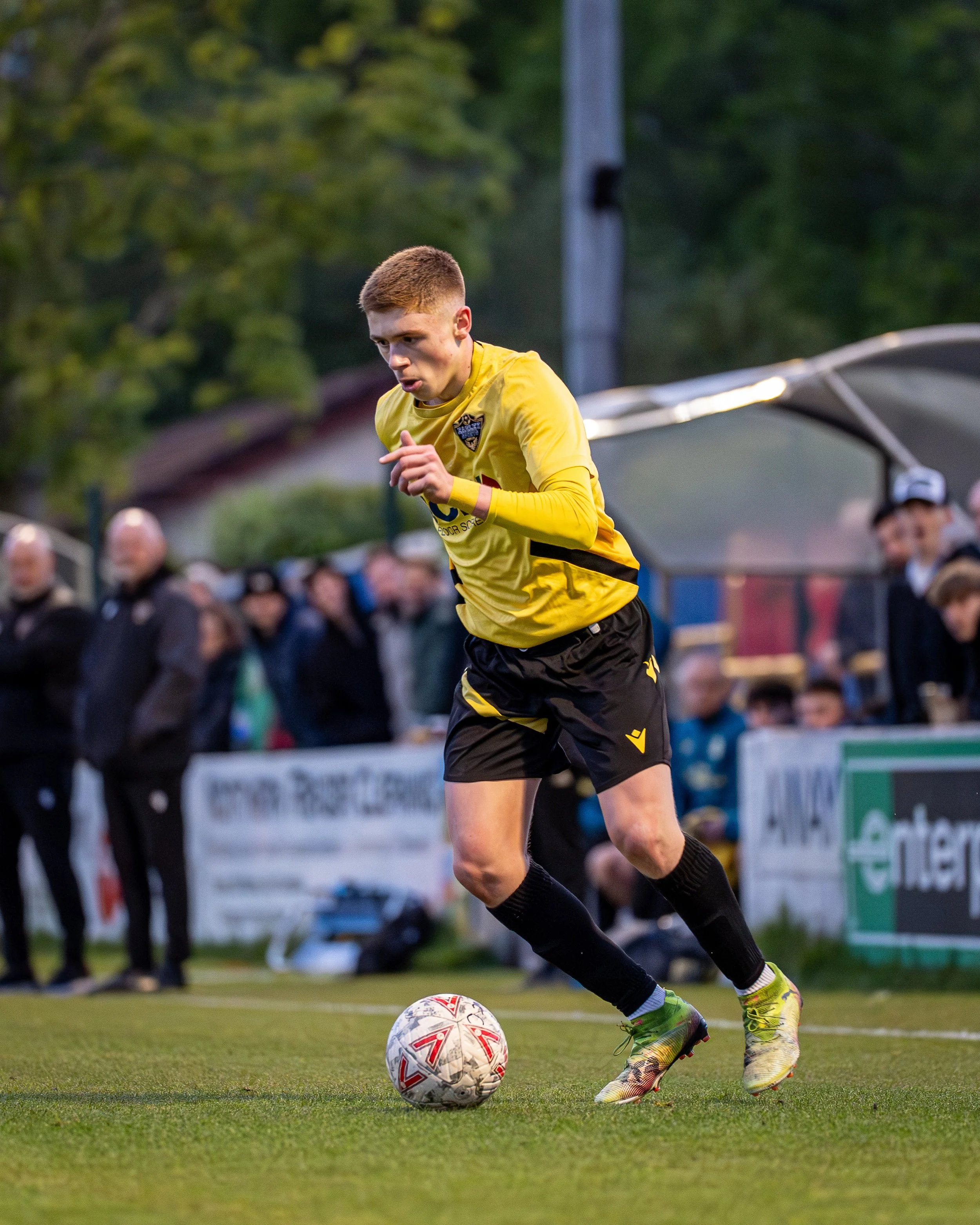 A male soccer player in yellow jersey and black shorts is running on a grass field, controlling a soccer ball with his right foot during a game or practice. Spectators are visible in the blurred background.