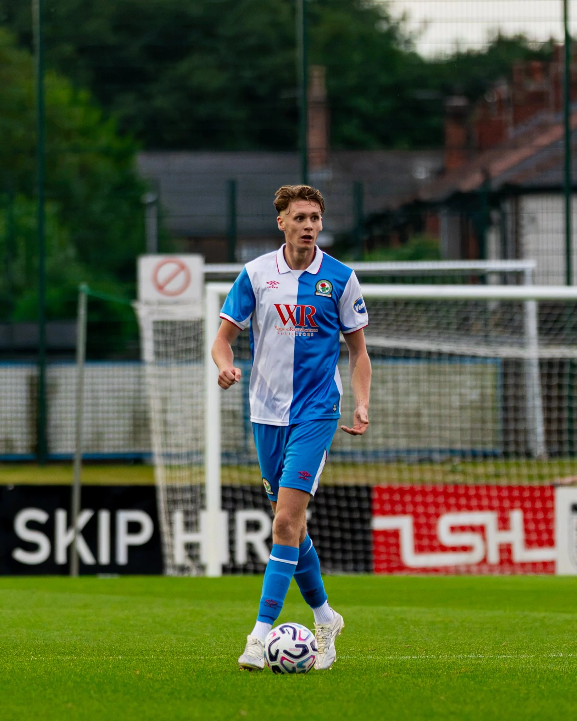 A young male soccer player in a blue and white uniform is standing on a grass field with one foot on a soccer ball. He appears to be preparing to kick, with a focused expression. In the background, there is a fence and some advertising boards.