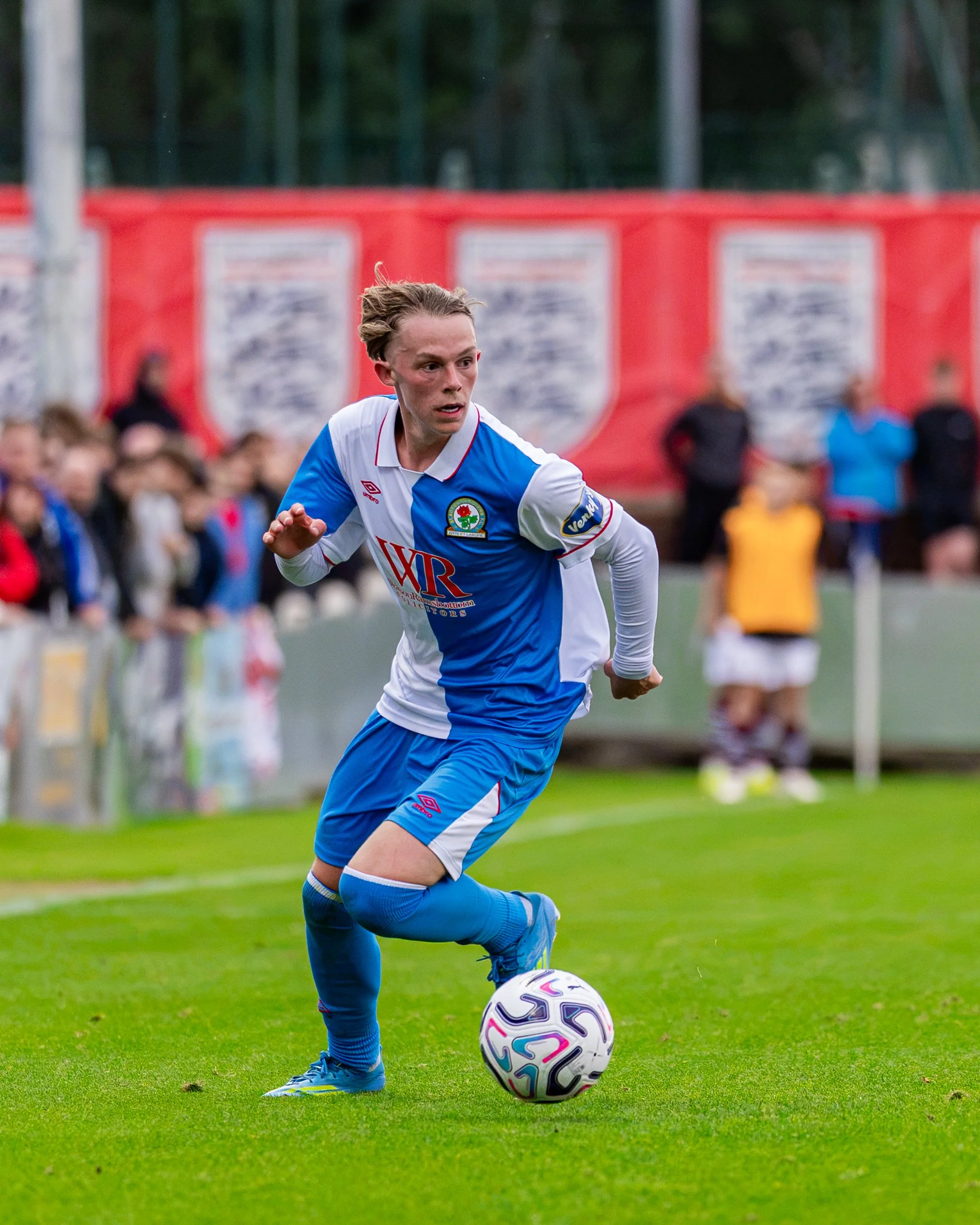 Soccer player in blue and white uniform kicking the ball on a green field, with an audience and red banners in the background.