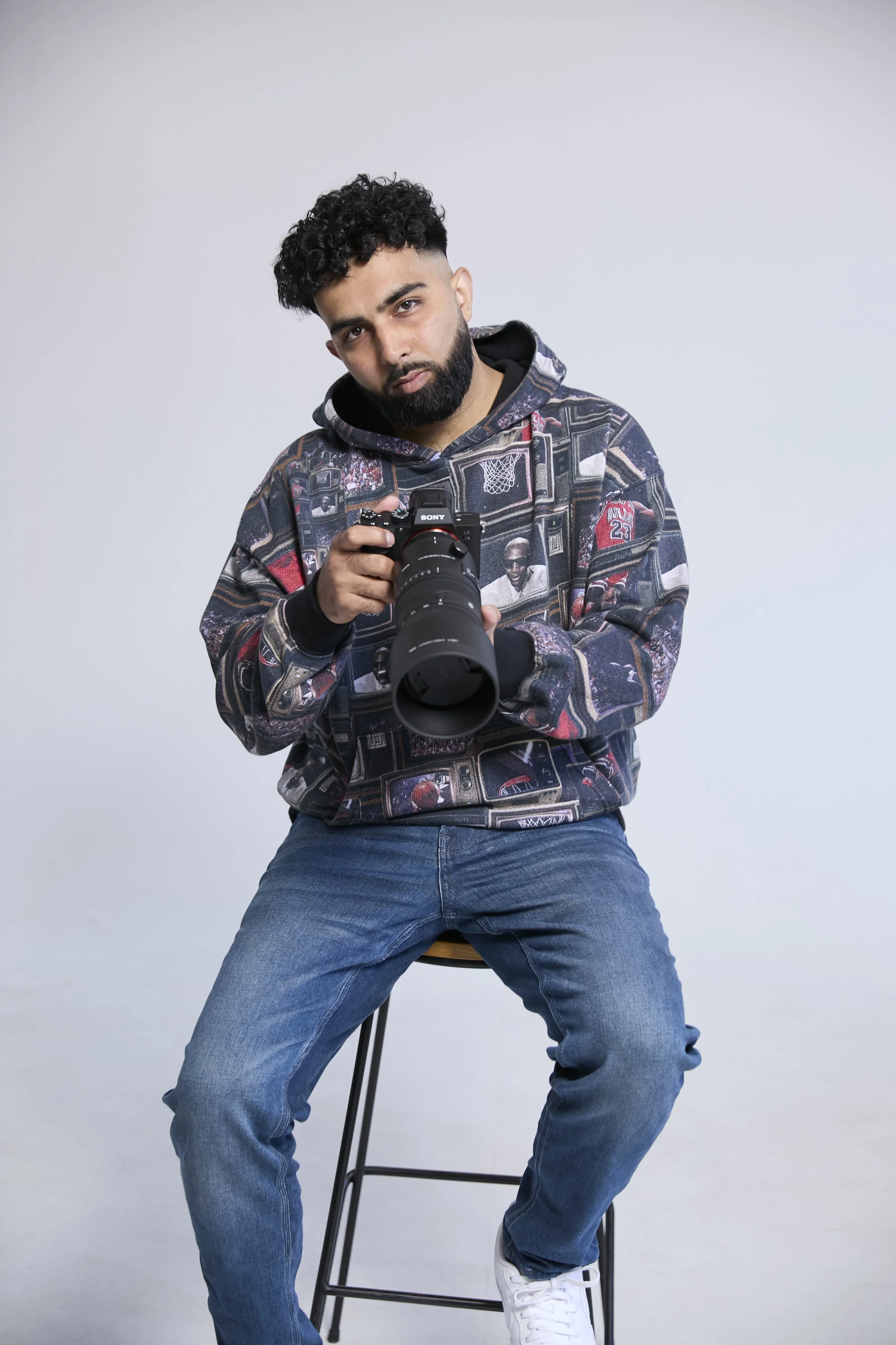 Young man with curly hair and beard sitting on a stool against a plain gray background, holding a camera and looking at the camera with a serious expression.