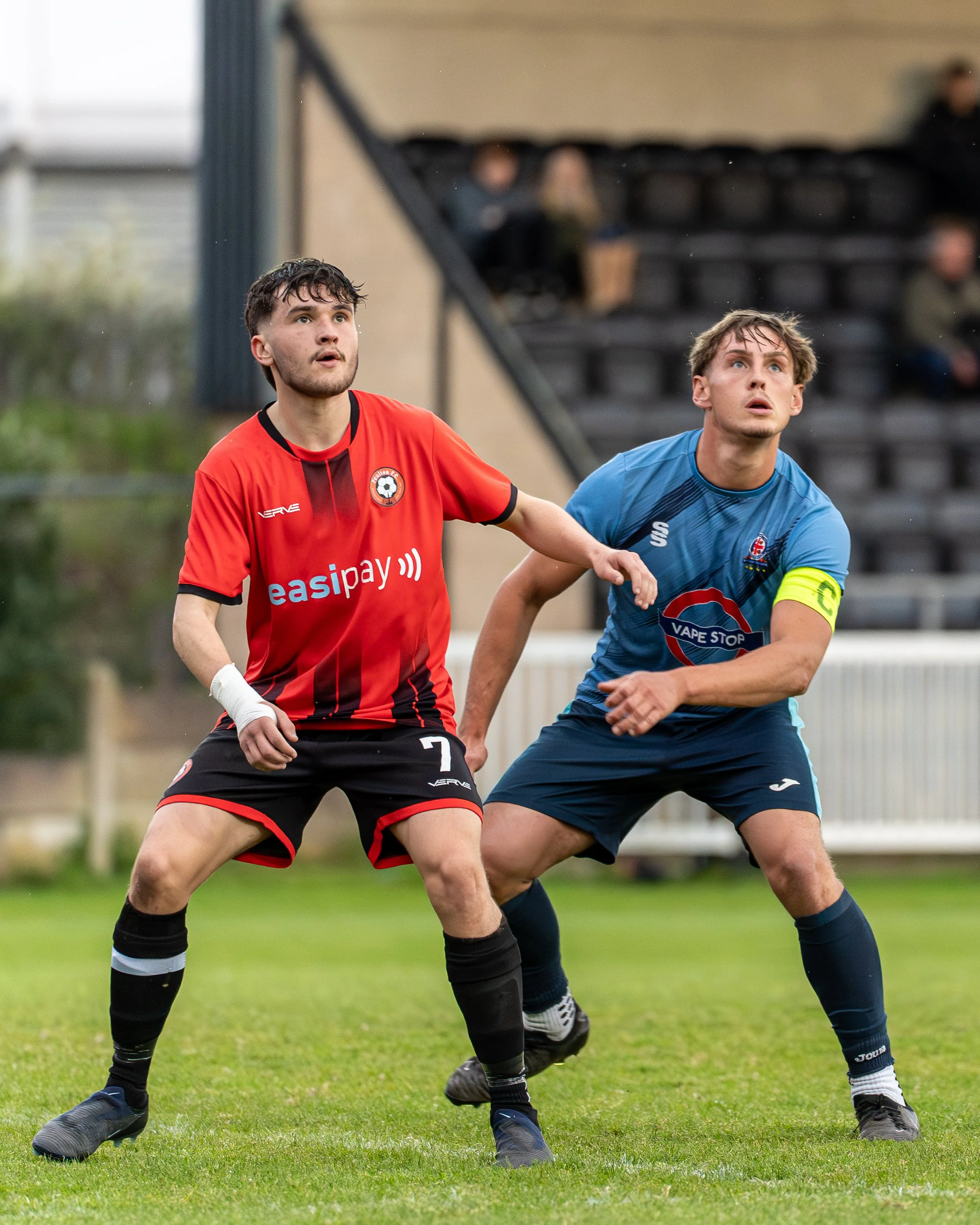 Two male soccer players are positioned on the field, looking upward, with a blurred background of seats and spectators.