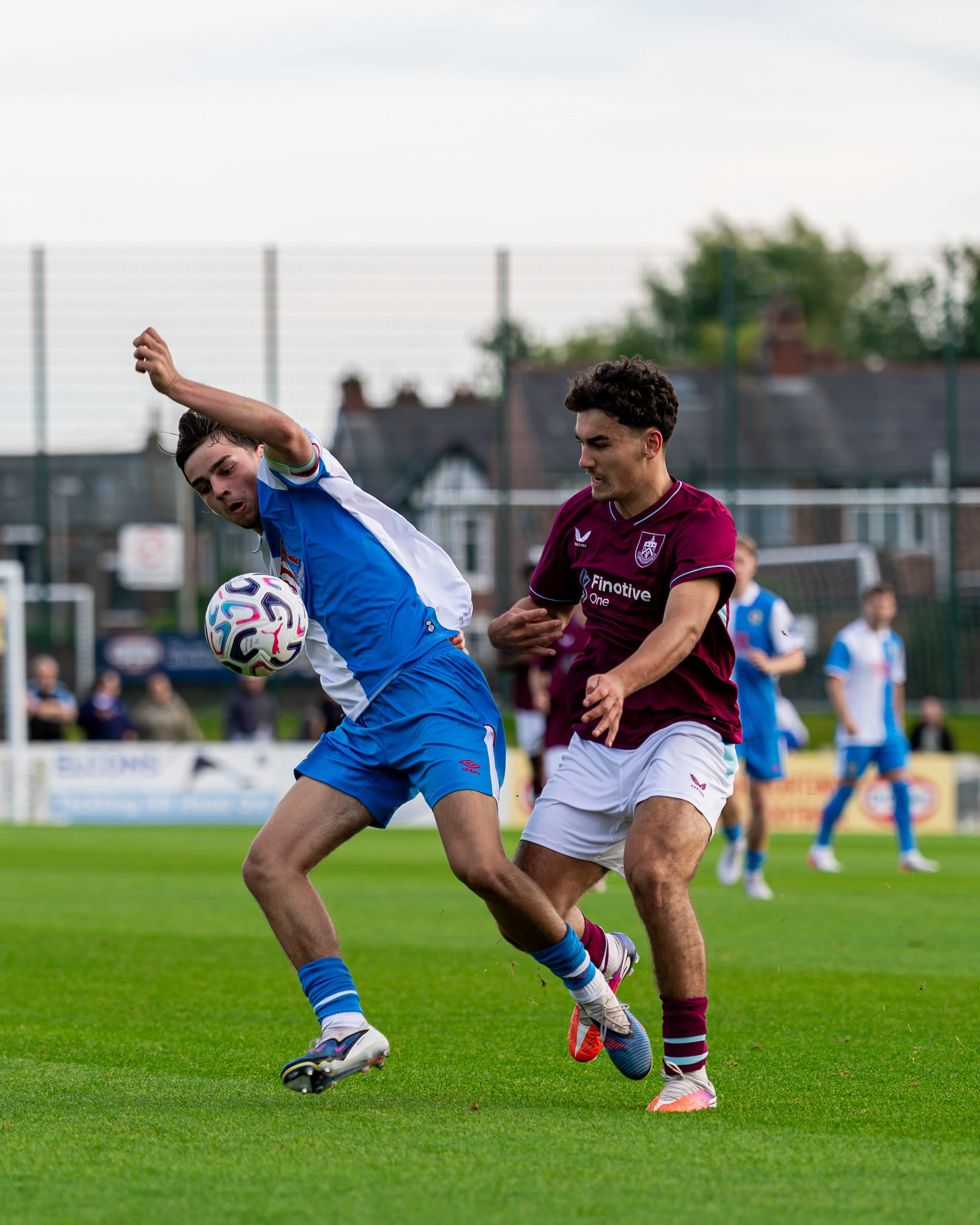 Two soccer players compete for the ball on a grass field, surrounded by other players and a blurred sideline scene.