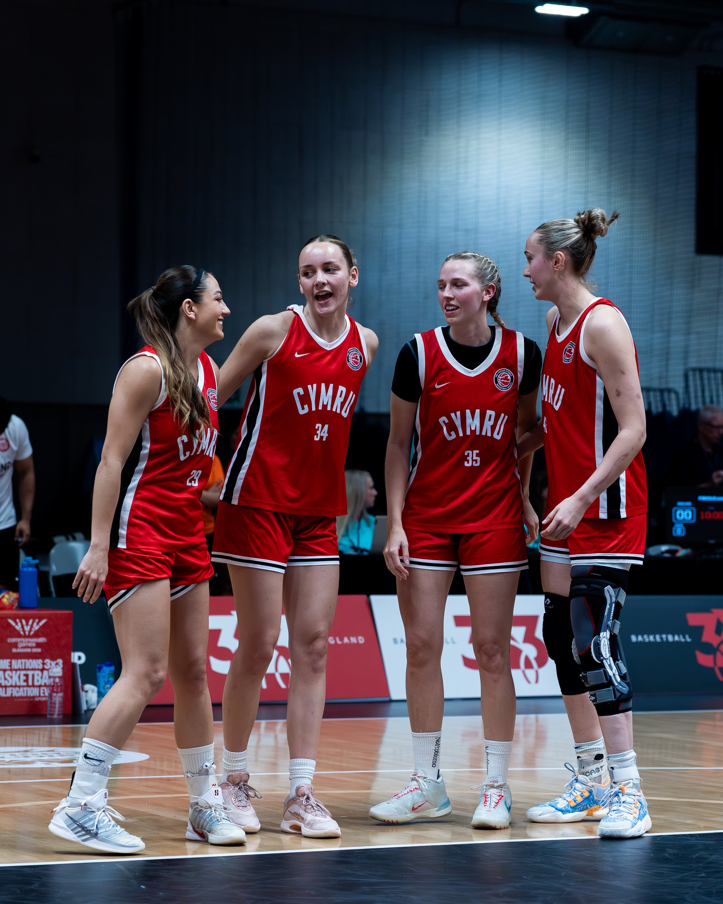 Four female basketball players in red jerseys standing together on a court, engaged in conversation during a game.