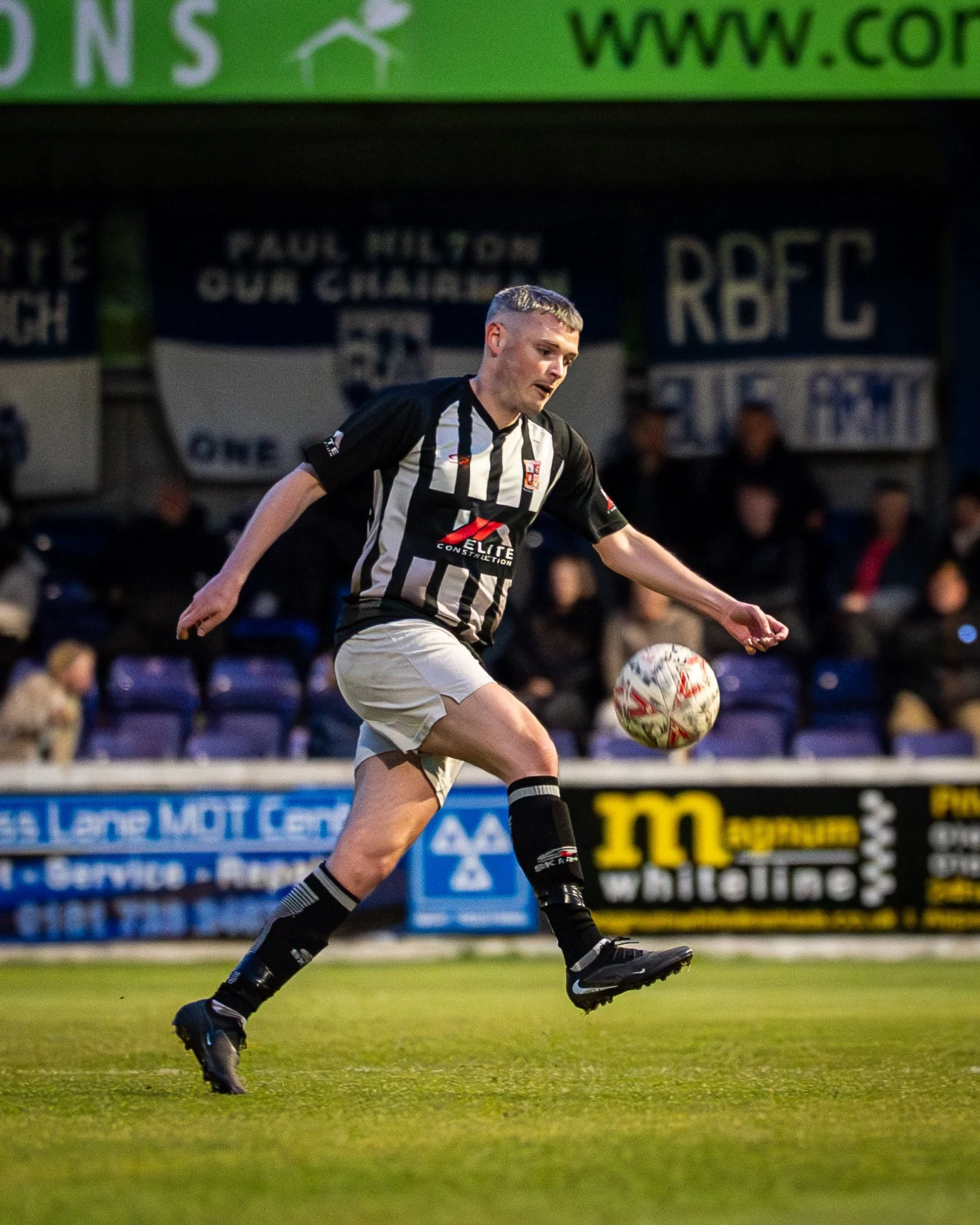 A soccer player in a black and white striped jersey kicks a soccer ball on the field during a game. The background shows a stadium with spectators and banners.