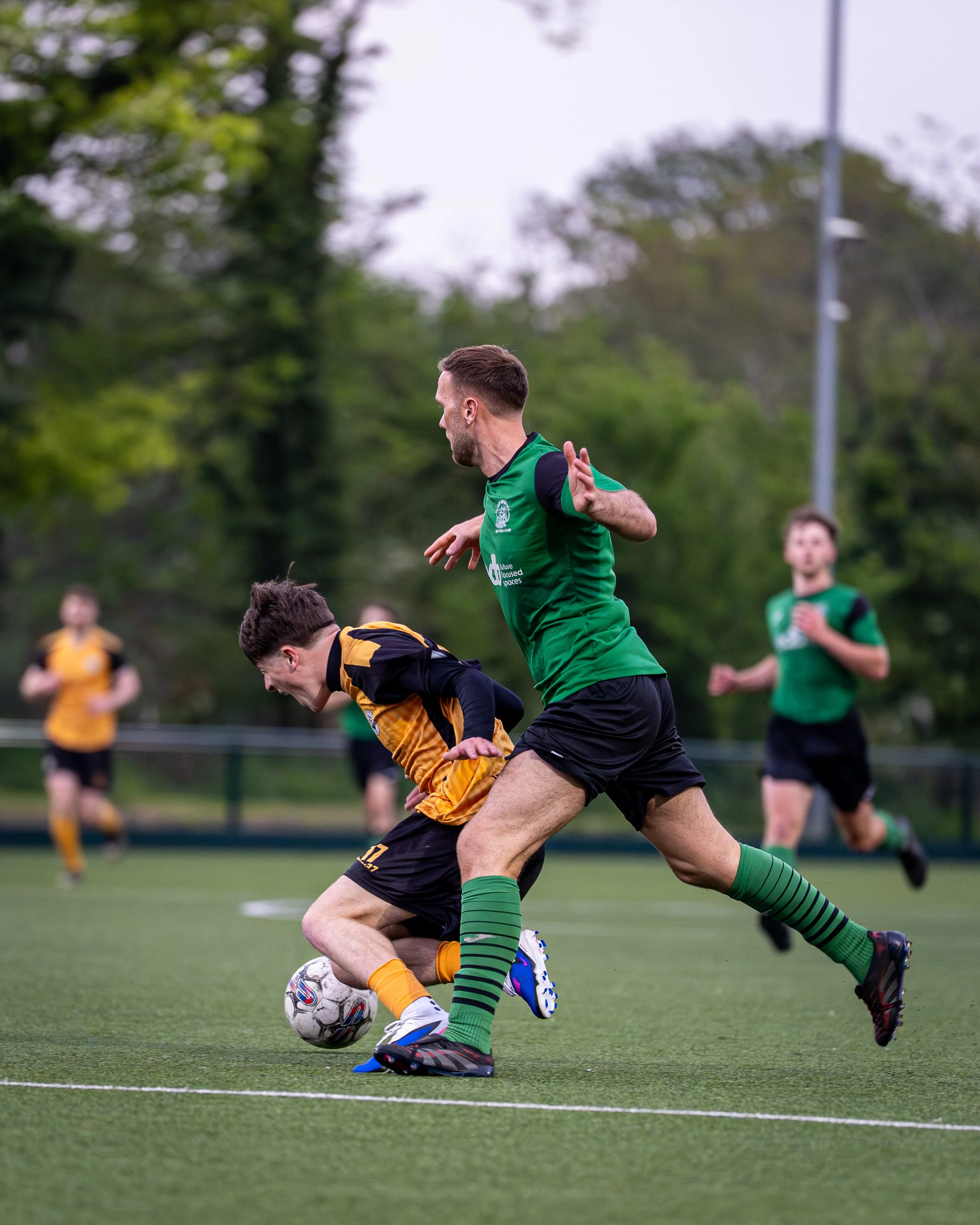 Two soccer players competing for ball, one in green jersey, black shorts, and striped green socks, the other in yellow and black jersey with black shorts, on a green field with trees and spectators in the background.