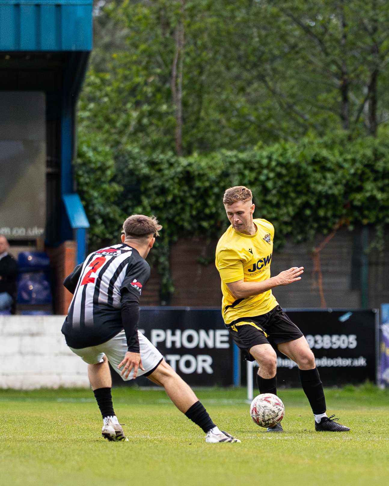 Two soccer players compete for the ball on a grass field, with one in a black and grey striped jersey and the other in a yellow jersey. There is greenery and advertising boards in the background.