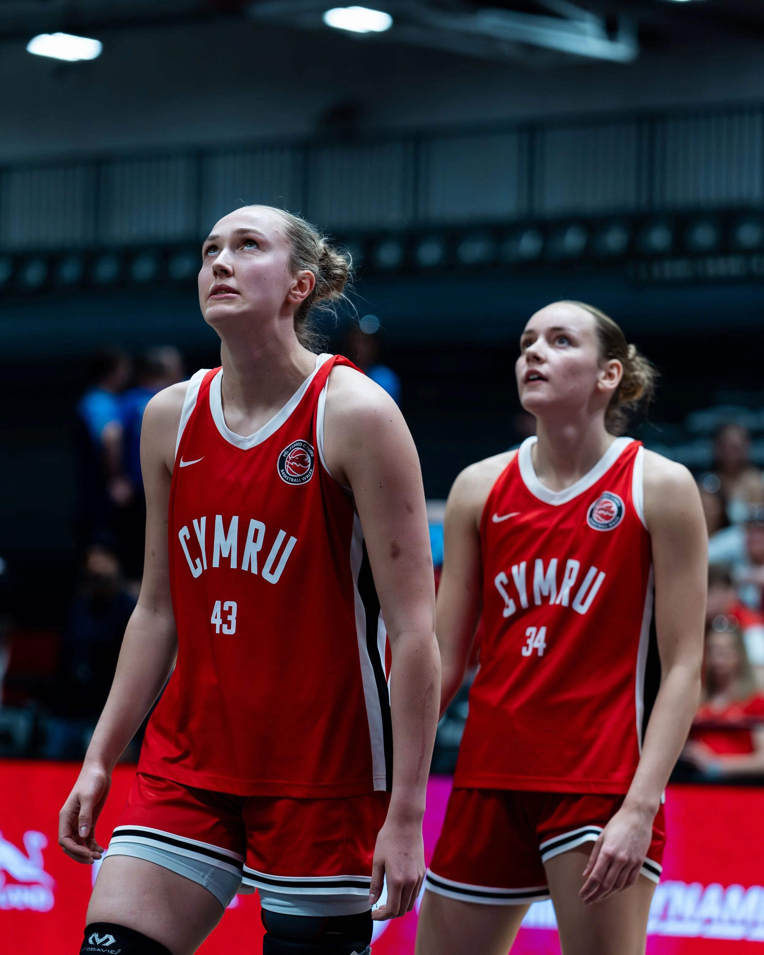 Two female volleyball players in red uniforms with white and black stripes, standing on a volleyball court, looking upward, with a dark background and lights.