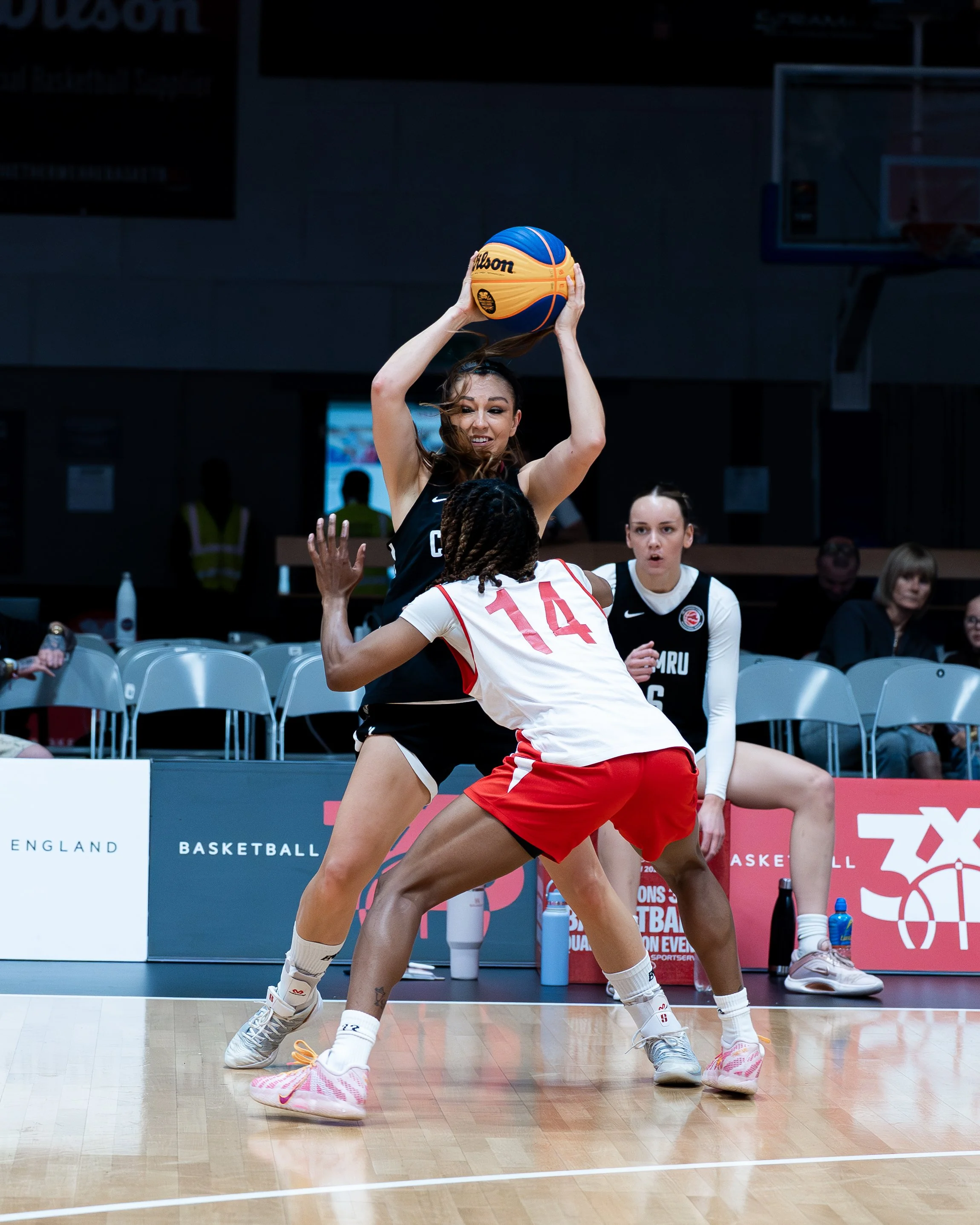 Two female basketball players are actively contesting for possession of the basketball on the court. One player with a dark jersey is reaching up to grab the ball, while the other with a white jersey numbered 14 is defending. The background shows a f