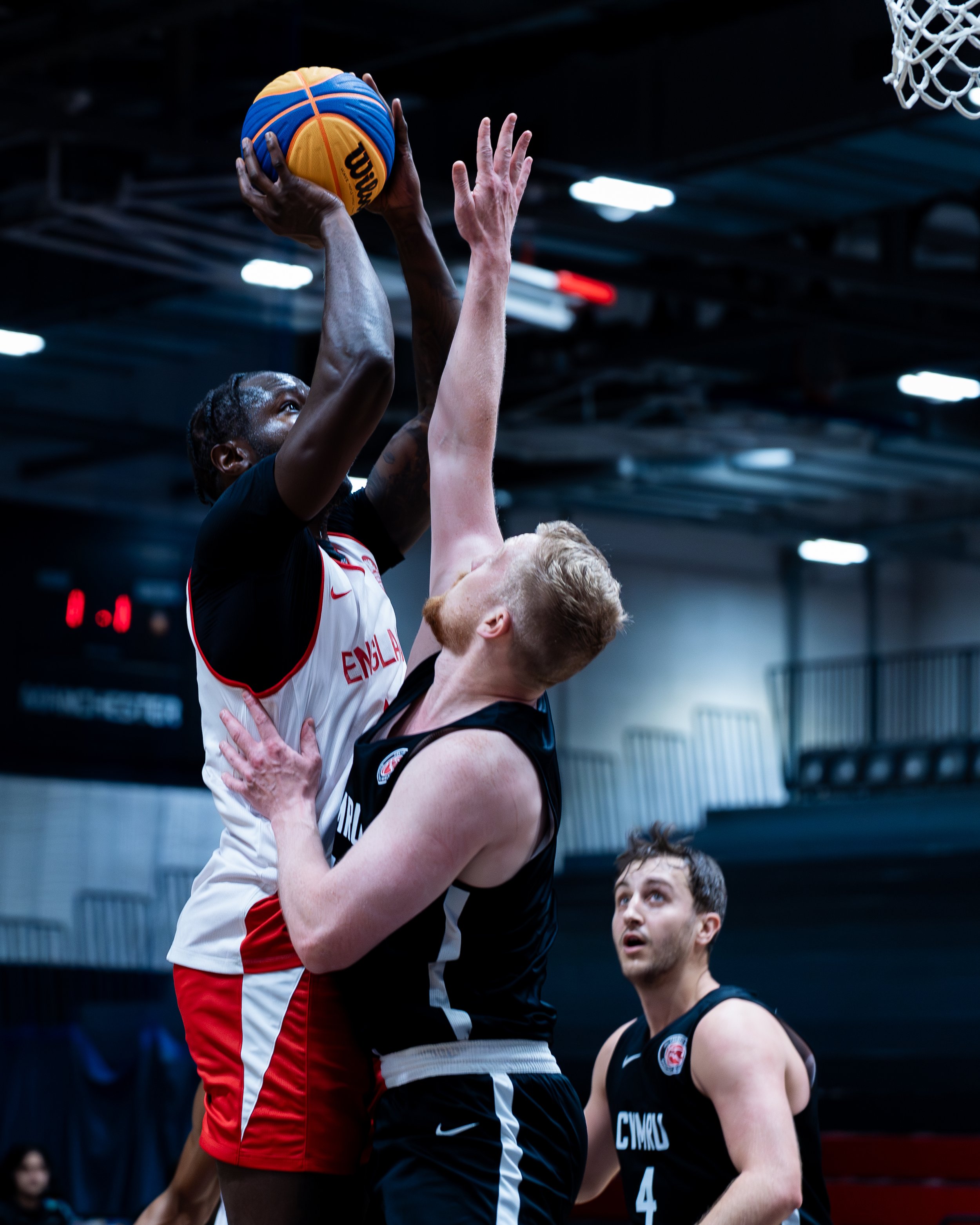 A basketball player in a white and red jersey jumps to shoot the basketball while an opponent in a black jersey tries to block the shot. The game is taking place indoors, with a dark ceiling and bright lights above.