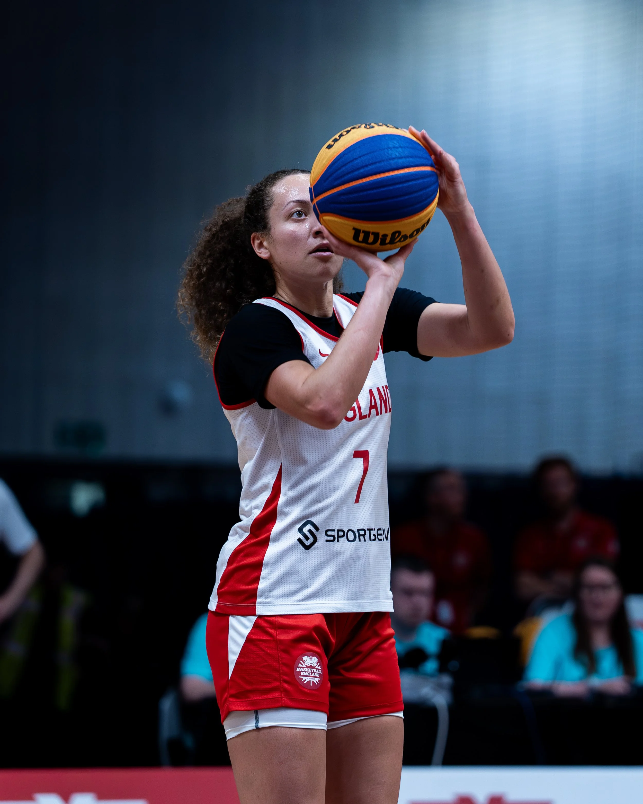A female volleyball player with curly hair taking a shot with a blue and yellow Wilson volleyball, in a sports arena with spectators in the background.