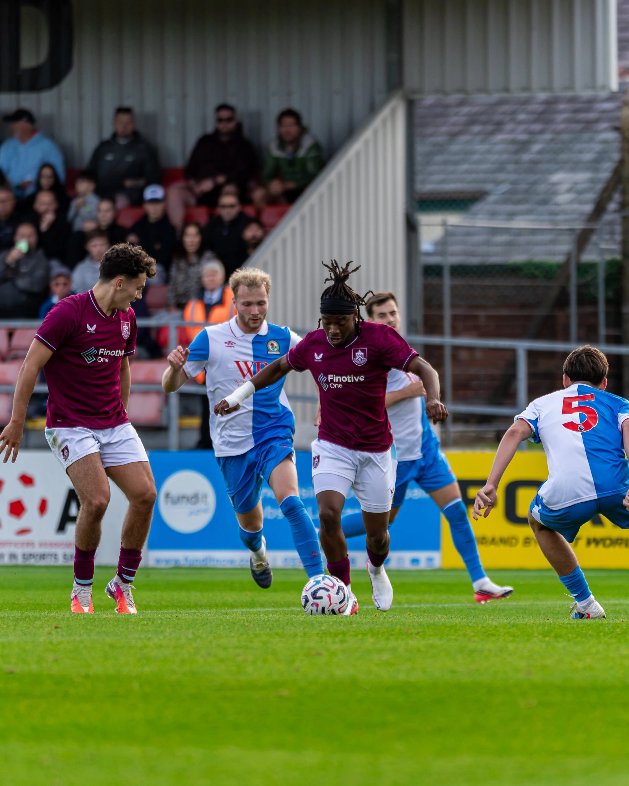 Soccer players competing for the ball on a field, with spectators watching from the stands in the background.