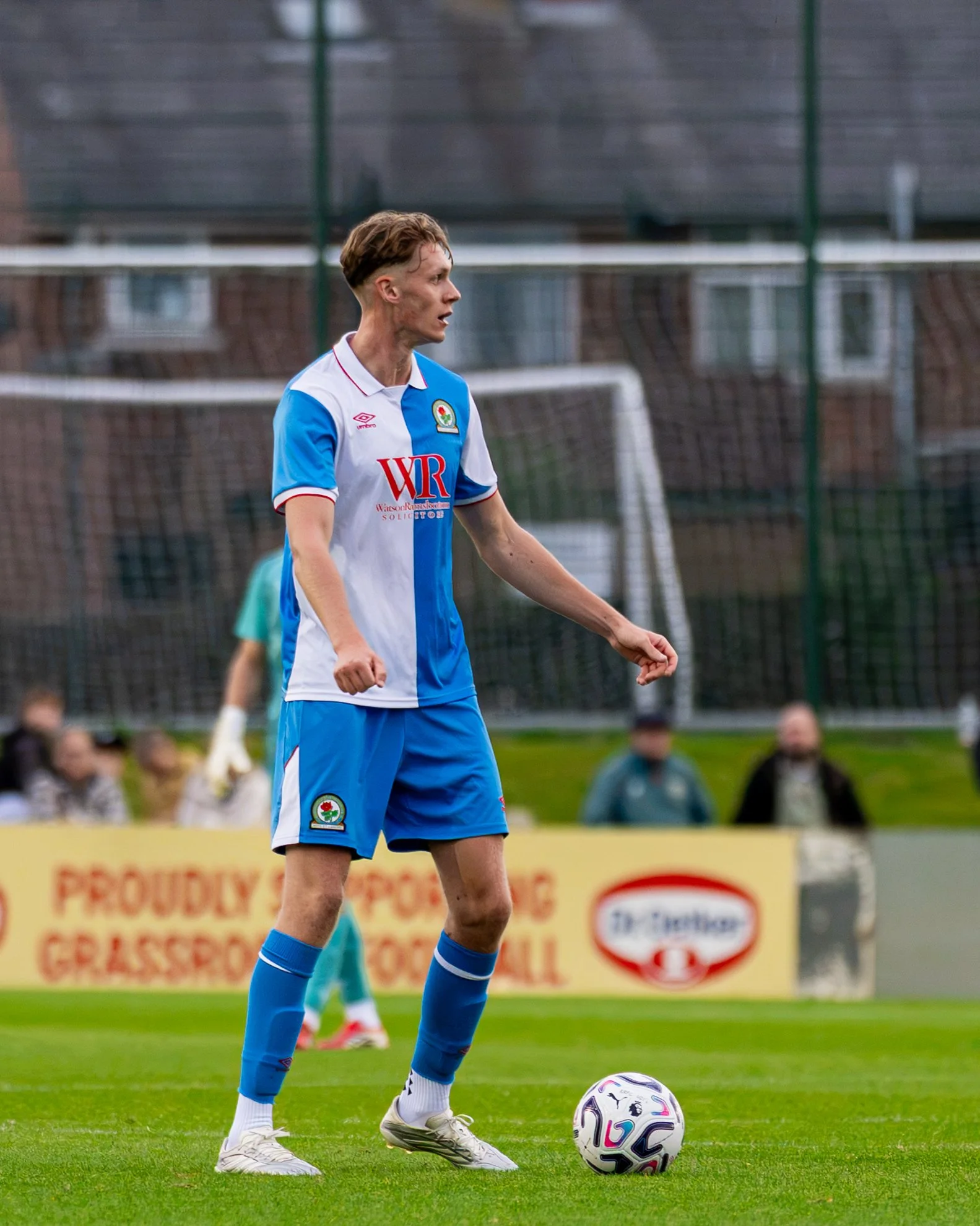 A male soccer player wearing a white and blue jersey with a logo, standing on a green field with a soccer ball at his feet, preparing to kick the ball against a background of a goal and blurred spectators.