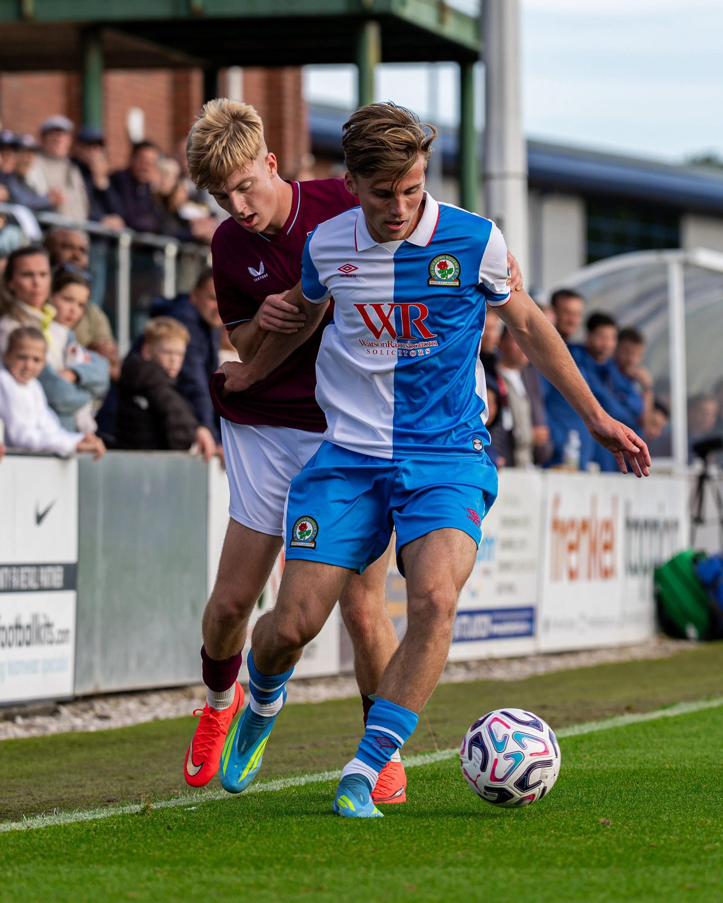 Two soccer players compete for the ball on the field during a match, with spectators watching from the sidelines. One player wears a maroon jersey and the other a white and blue jersey with a rose emblem.