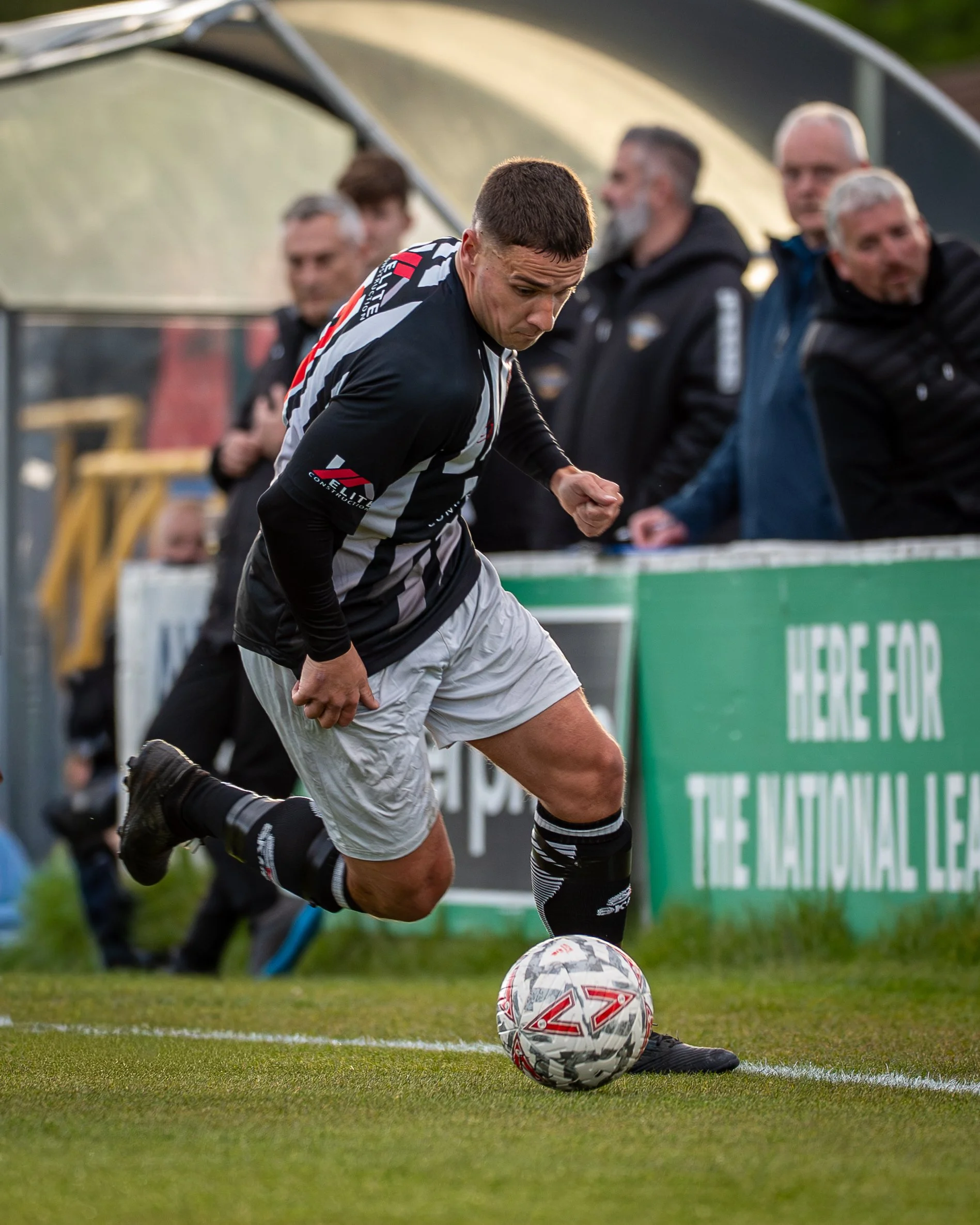 A soccer player in black and white uniform kicking a soccer ball on the field, with spectators and advertising boards in the background.