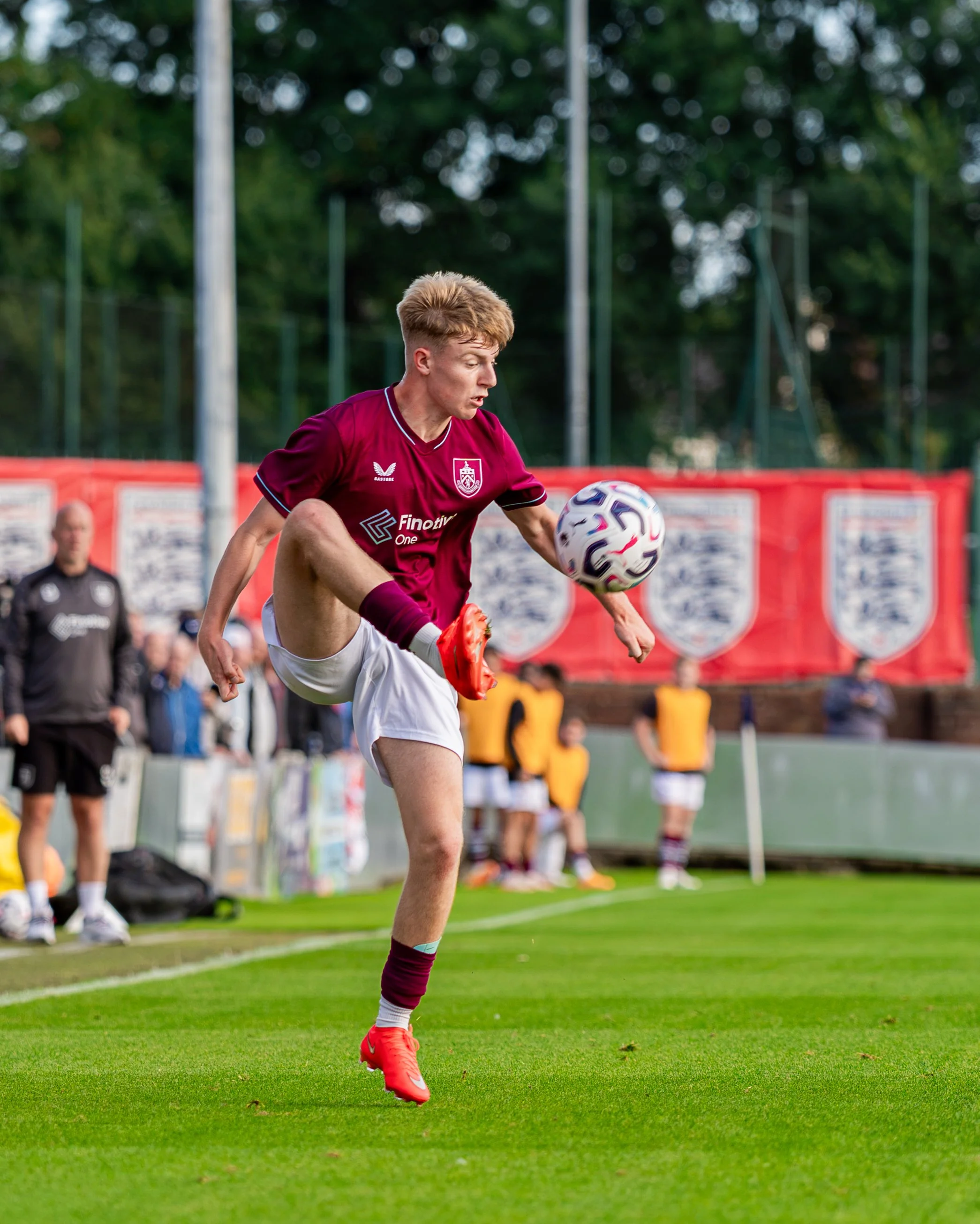 A soccer player in a maroon uniform and orange cleats kicks a soccer ball on a field during a match, with a group of spectators and a referee in the background.