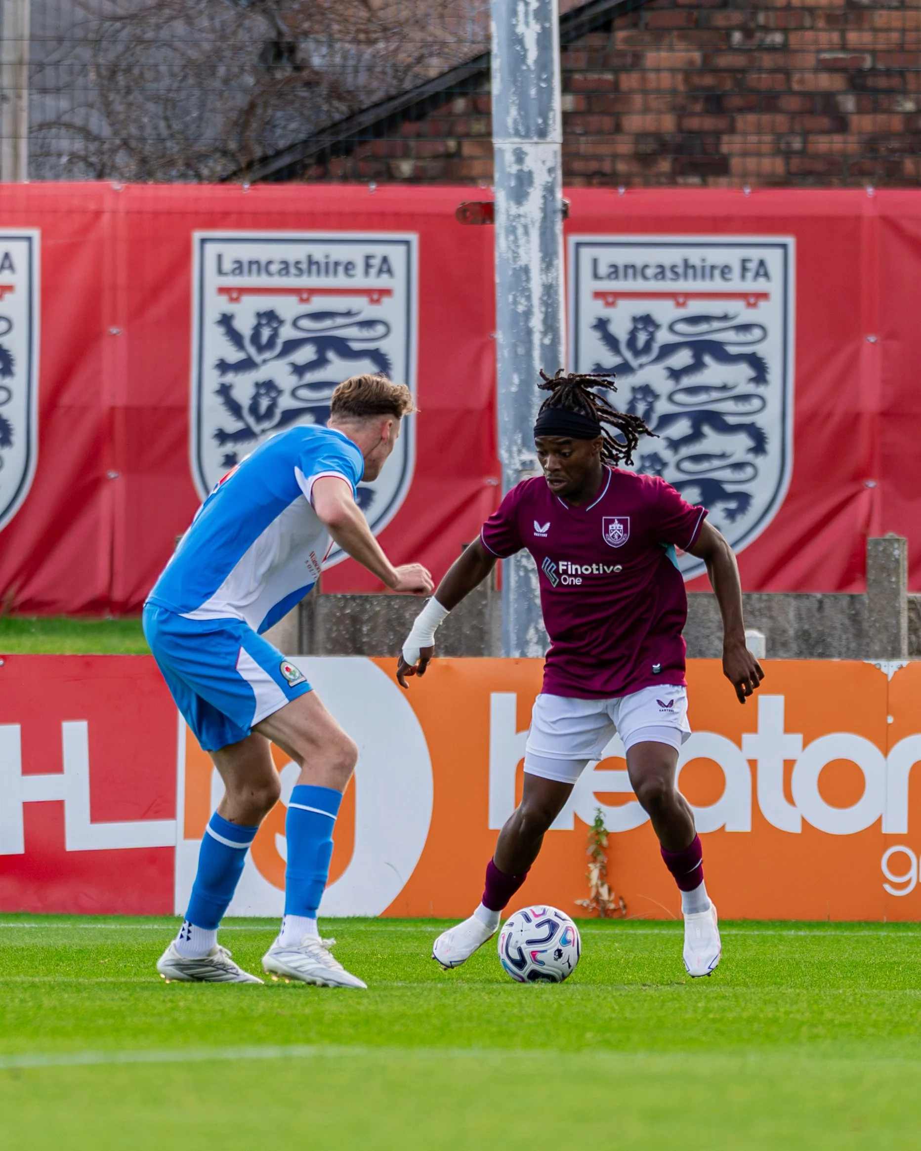 Two soccer players compete for the ball on the field during a match, with a red banner displaying the Lancashire FA logo in the background.