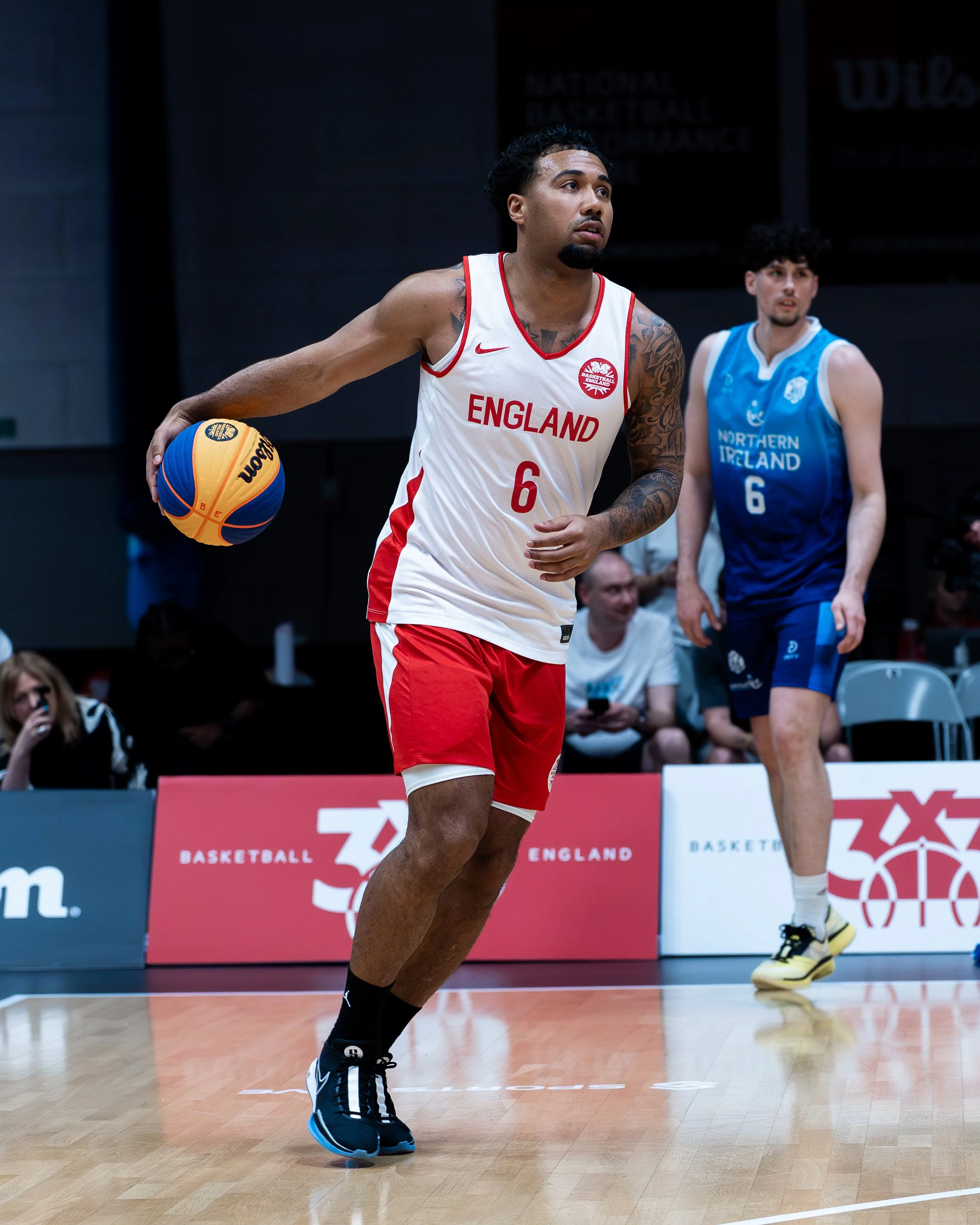 A basketball player in a white and red England jersey with the number 6 dribbling a basketball on an indoor court, with another player in a blue Ireland jersey in the background.