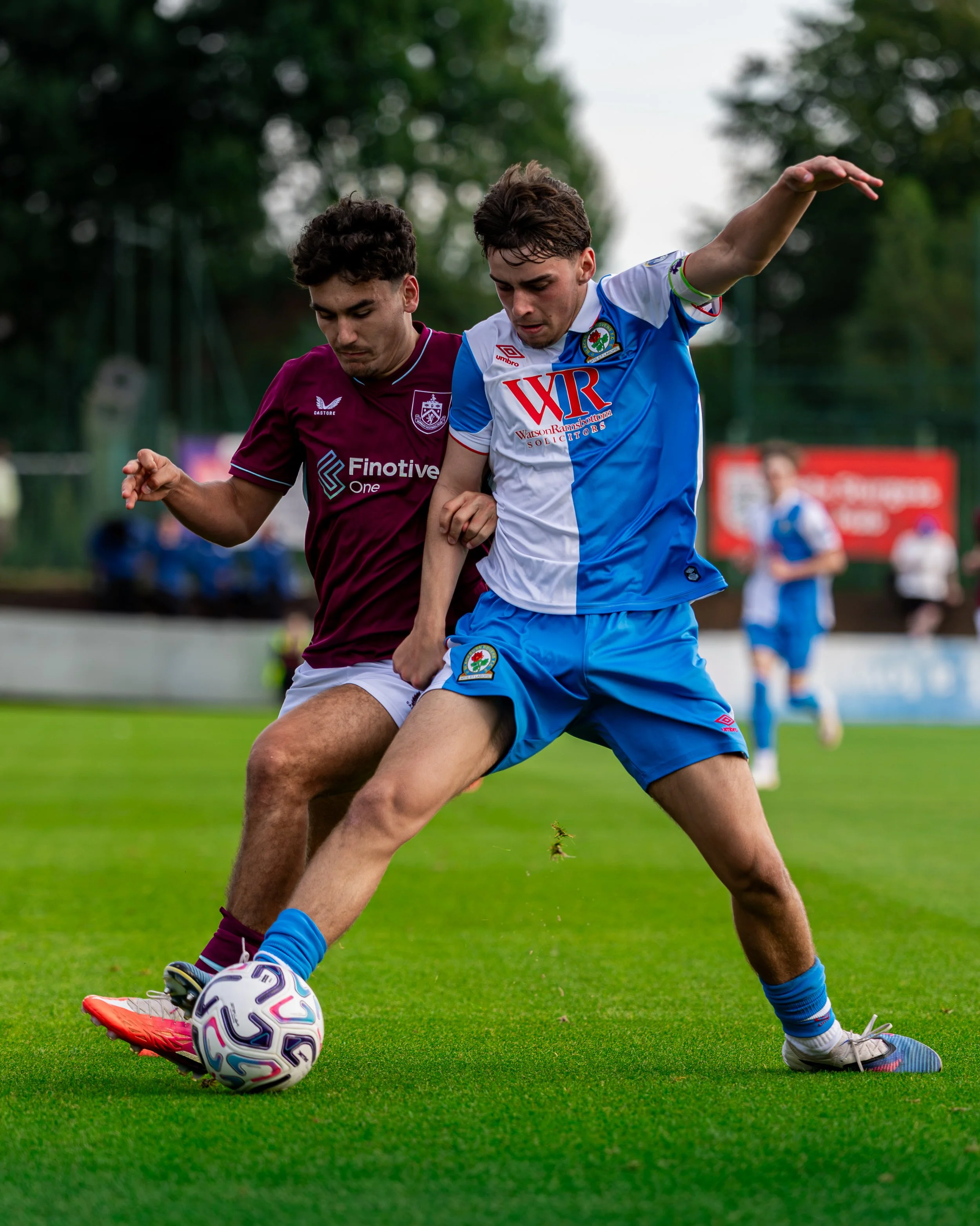 Two soccer players competing for the ball on a grass field, with trees and blurred players in the background.