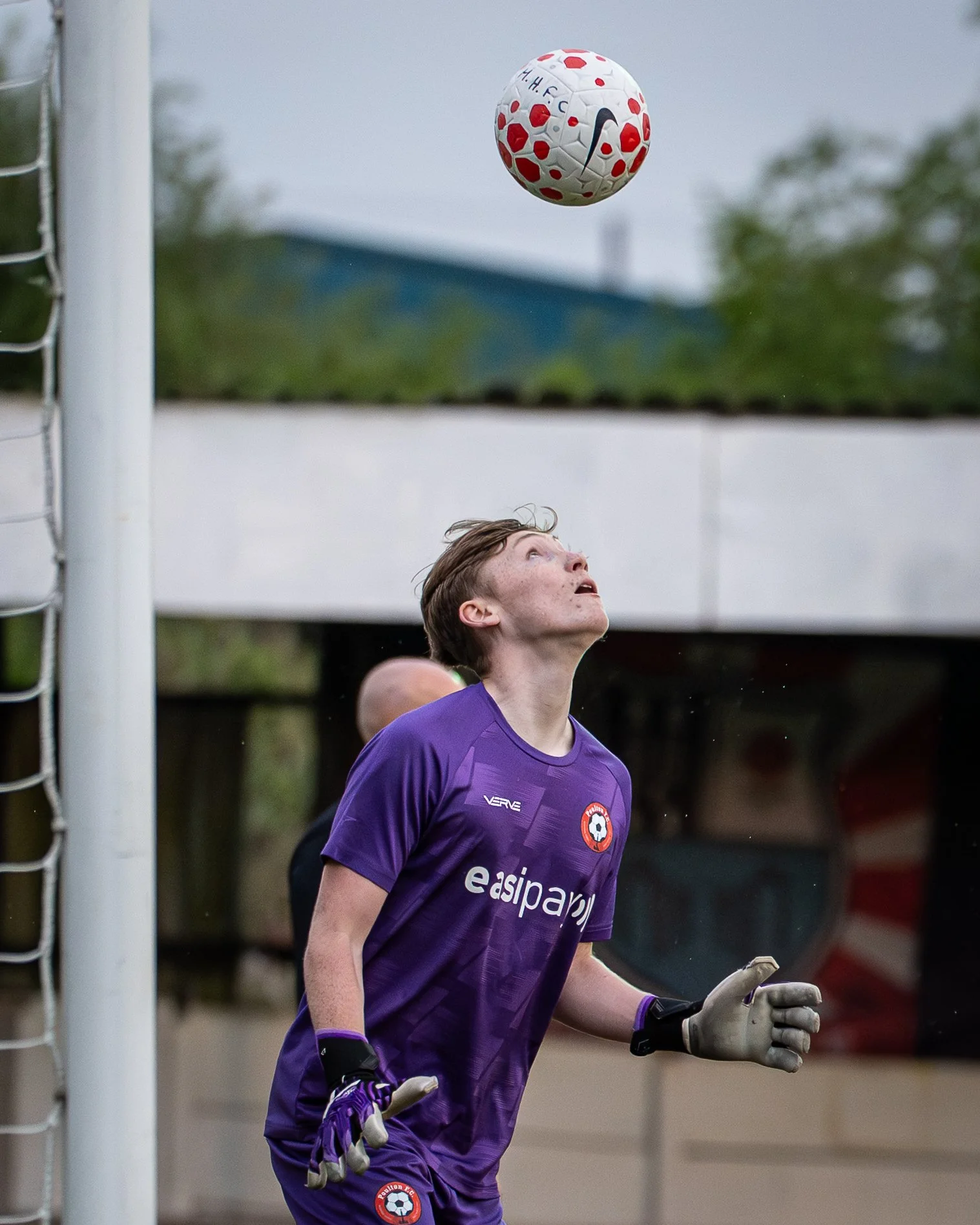 A soccer goalkeeper in a purple jersey with a logo and the word 'easpay' on it, wearing gloves, looking upward as a soccer ball with red and black markings flies towards the goal.