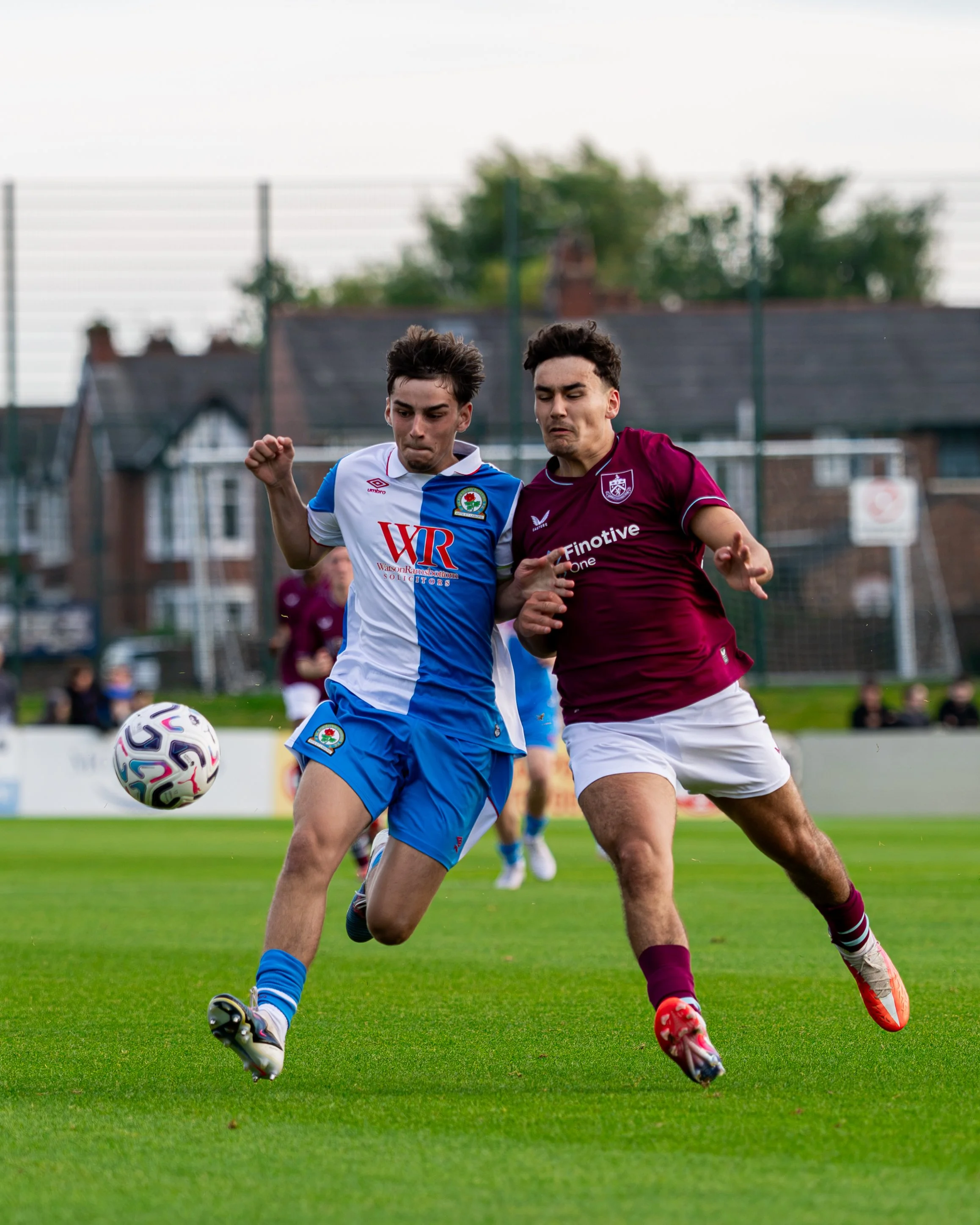 Two male soccer players competing for the ball during a match on a grass field, with one player in a blue and white kit and the other in a maroon kit, both focused on the game.