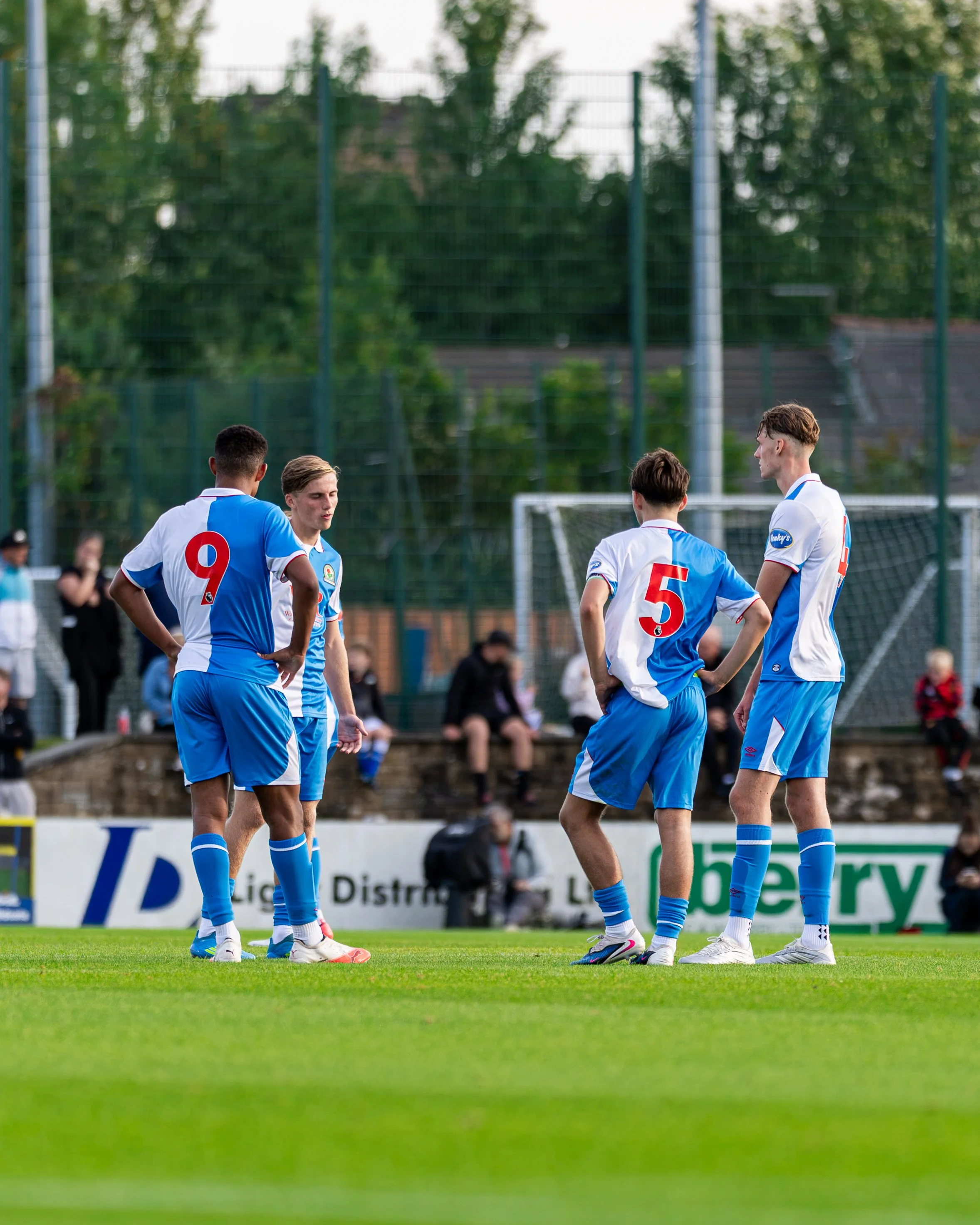 Four soccer players wearing white and blue uniforms are standing on a green soccer field, seemingly engaged in a discussion or strategy during a match. Spectators are in the background along the sidelines.