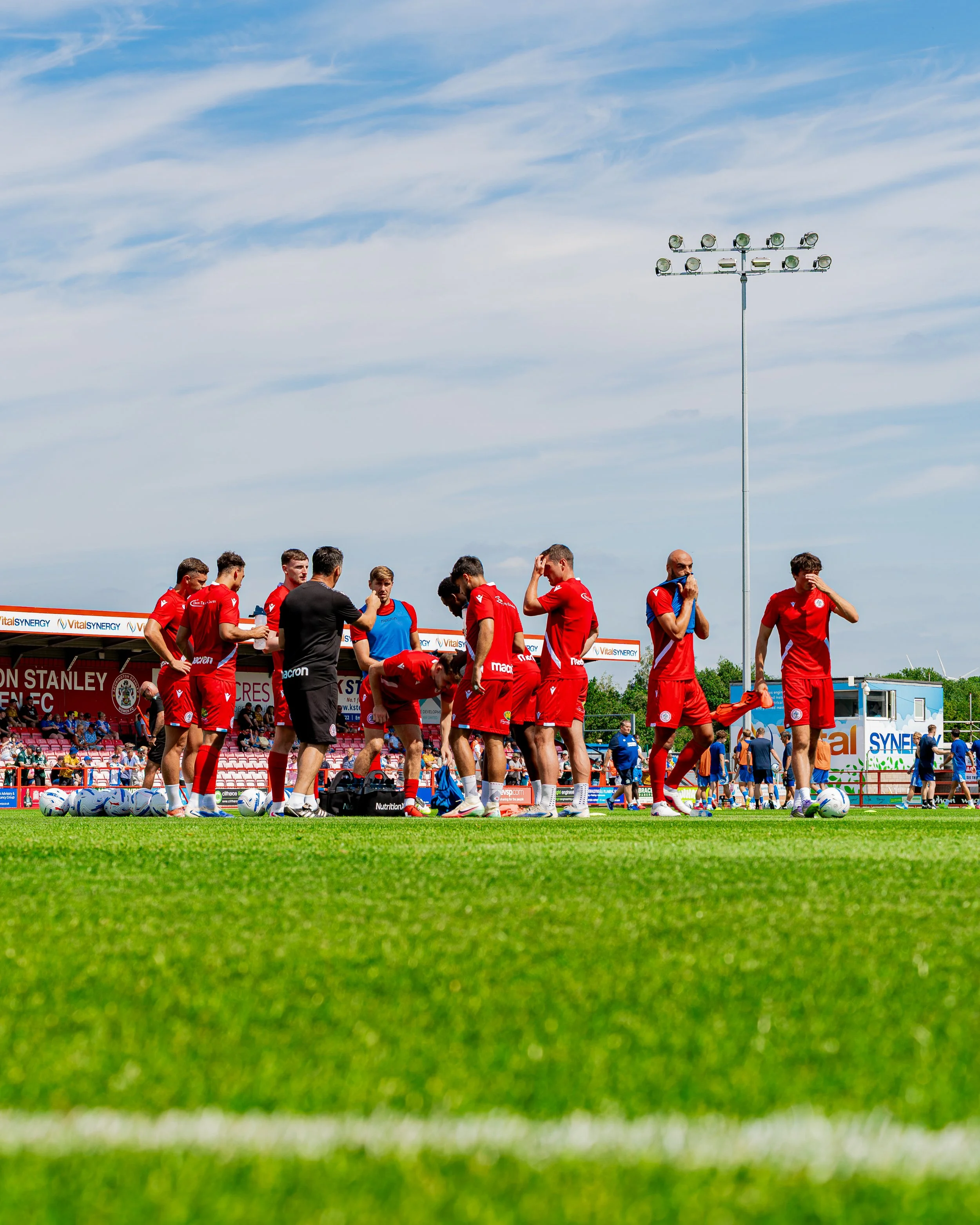 Soccer players in red jerseys are on a field with footballs, preparing for a match or practice, with a blue sky and stadium in the background.