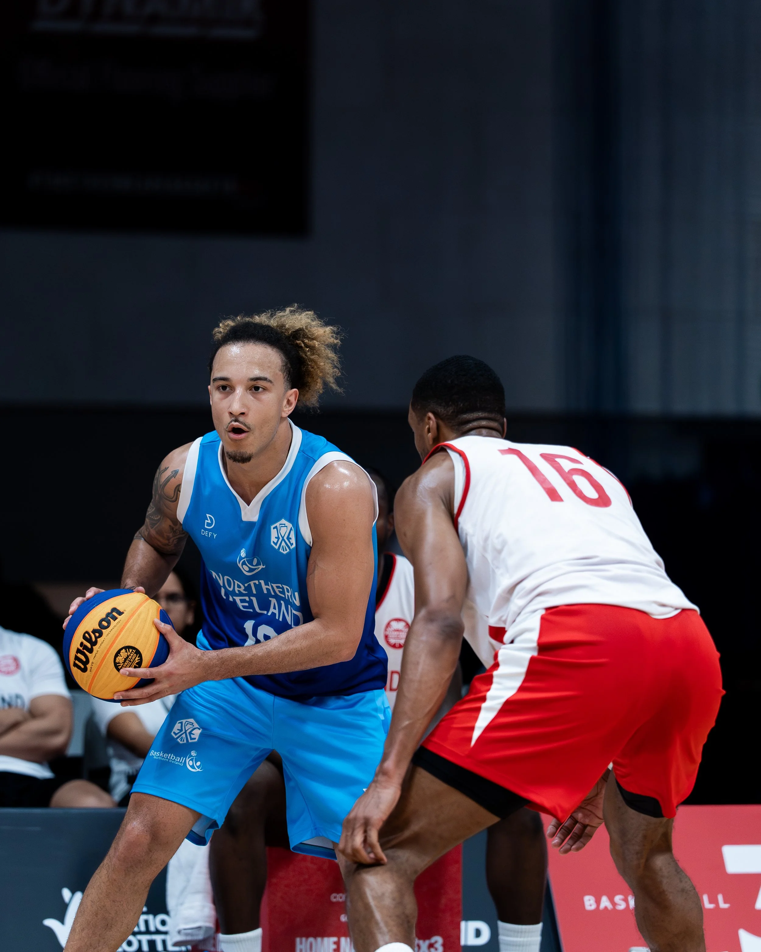 Two basketball players, one in a blue uniform holding a basketball and another in a white and red uniform opponent.
