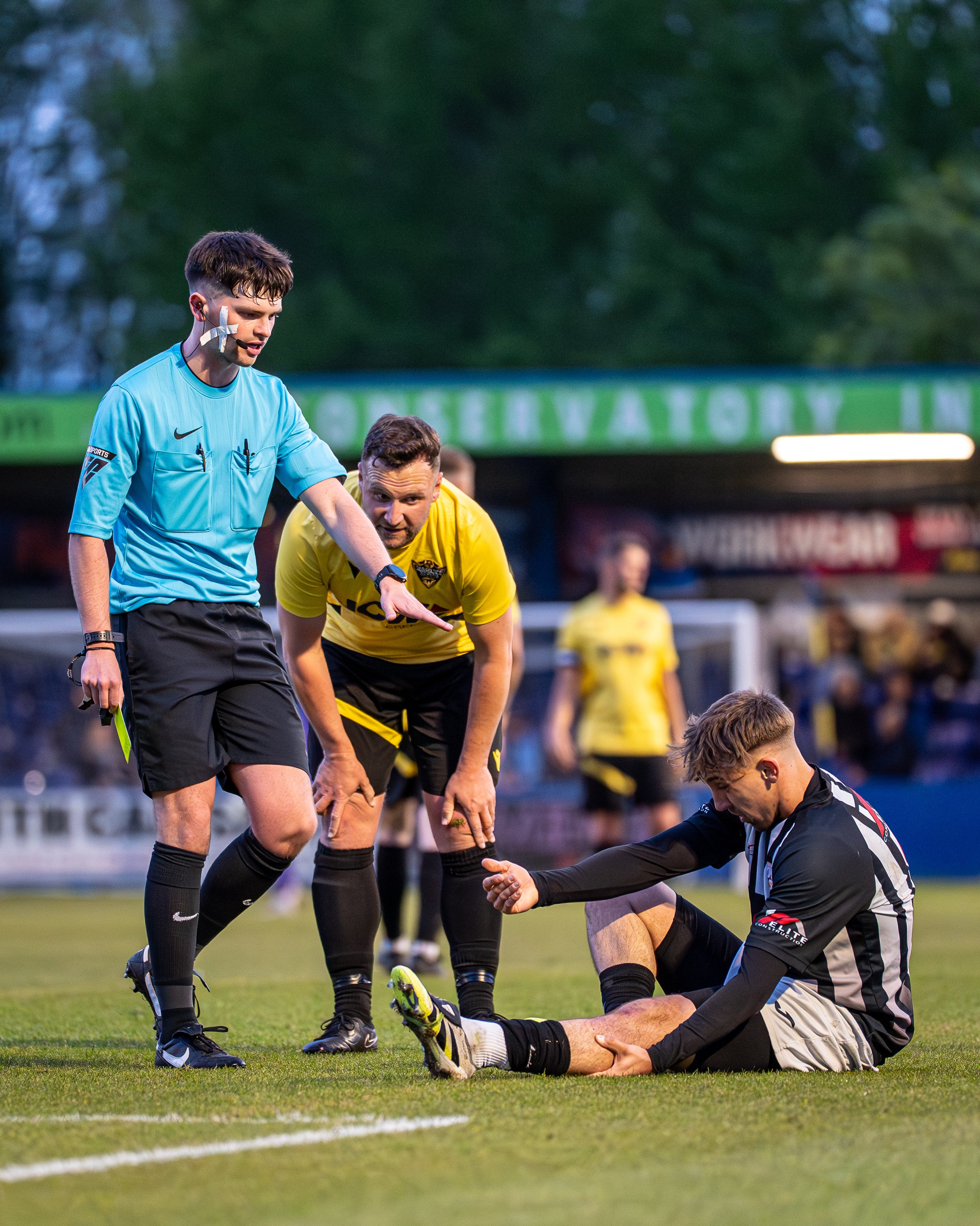 A soccer match scene with a referee and players. One player is sitting on the ground receiving medical attention from a teammate, while the referee looks on. The background shows a blurred crowd and trees.